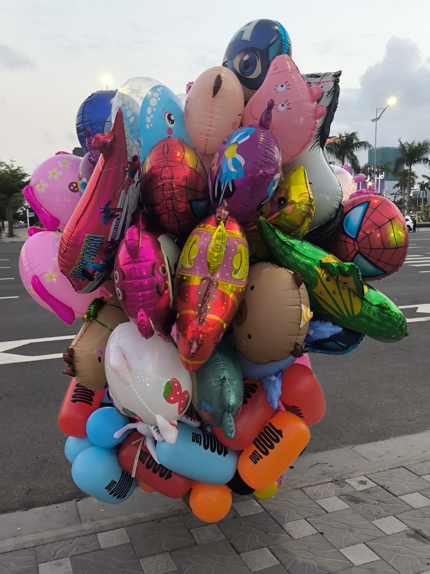 A large, dense cluster of approximately 30-40 inflated foil and plastic balloons dominates the center and foreground of the image, appearing to be held just above a sidewalk. The balloons vary widely in shape, color, and character, including representations of popular culture figures such as Spiderman (red with web pattern, and on an elongated pink balloon) and Captain America (a blue shield-like balloon), along with various animals and cartoon characters like a pink fish, a green crocodile, a blue whale/jellyfish, a brown bear-like head, a white bunny/pig with a strawberry motif, a pink star-shaped creature, and a white horse head. Generic shapes include pink oval balloons with floral patterns, elongated pink, red, and yellow balloons, and cylindrical red and blue balloons marked with "1000 ton" in black text. The balloons feature vibrant colors such as pink, red, blue, green, purple, yellow, orange, white, and brown.
The balloons are situated on the edge of a sidewalk composed of gray and white checkered paving stones, which borders an asphalt road. White road markings, including a forward arrow and solid lines, are visible on the asphalt. In the background, a line of palm trees and other tropical foliage is present. Modern buildings, some partially obscured and featuring illuminated elements, rise behind the trees. A bright street light on a tall pole is visible on the right, emitting a yellow glow against a light gray, overcast sky, indicative of dusk or an overcast day. Small, indistinct figures and vehicles are visible on the distant road. The wide thoroughfare, tropical vegetation, and modern urban infrastructure are characteristic of coastal areas in Đà Nẵng, Vietnam, likely near a major boulevard such as those running parallel to the beaches.