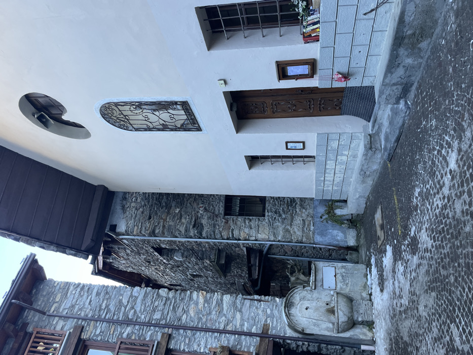 The image captures a narrow, traditional street or alleyway in Courmayeur, Italy, characterized by rustic stone architecture and a contrasting lighter facade. The scene is quiet and appears to be daytime, with clear lighting.
On the left side, a multi-story building features a light-colored upper section with distinct architectural elements, transitioning to dark, rough-hewn stone masonry at its base. The upper part of this building includes a semi-circular cutout in the wall, possibly housing a speaker or light. Below this, an arched window is adorned with intricate black wrought-iron grilles. A dark brown wooden door with carved details serves as the main entrance, accessible via a small set of light-colored stone steps with a dark doormat. Above the door, a small rectangular window with an unreadable sign is present, and on the windowsill to its right, a stack of colorful books is visible. Below the door, another small framed sign displays what appears to be "HB" or similar text. Further down, a smaller window with simple grilles is set into the wall.
Opposite this building, on the right side of the alley, stands another structure made entirely of dark stone, which also has a small barred window. A metal pipe runs horizontally along its wall. In the foreground on the right, a traditional stone water fountain with a metal spout and a large basin is situated, adding to the historic ambiance. The ground is paved with small, dark, possibly cobblestone-like material, showing some lighter, wet patches or melted snow/ice. A small amount of greenery grows at the base of the lighter building. No people are visible, and no specific event is taking place, presenting a tranquil streetscape.