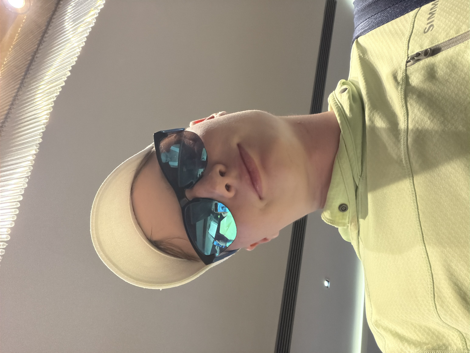 A young person wearing a light beige baseball cap and dark sunglasses is photographed from a low angle. They are also wearing a light green hooded jacket with a zipper. The sunglasses reflect two people and what appears to be a smartphone. The background shows a ceiling with decorative lighting fixtures and what looks like a ventilation system. The image does not provide any specific visual cues that would indicate the location is Kemer, Turkey.