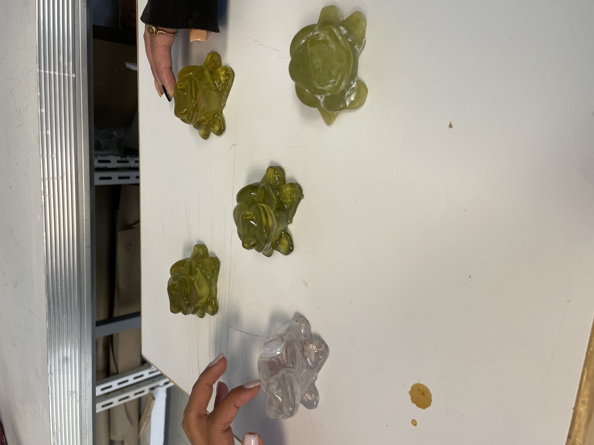 The image shows a white table in a room with a metal shelf in the background. Five small, translucent objects, resembling turtles, are scattered on the table. Four of these objects are light green, while one is clear. Two hands are visible; one with black nail polish touches one of the green objects, and the other with white nail polish touches the clear object. A small, dark spot is present on the table's surface. The location is not specified in the image, but the prompt states the image is from San Andrés Cholula, Mexico.