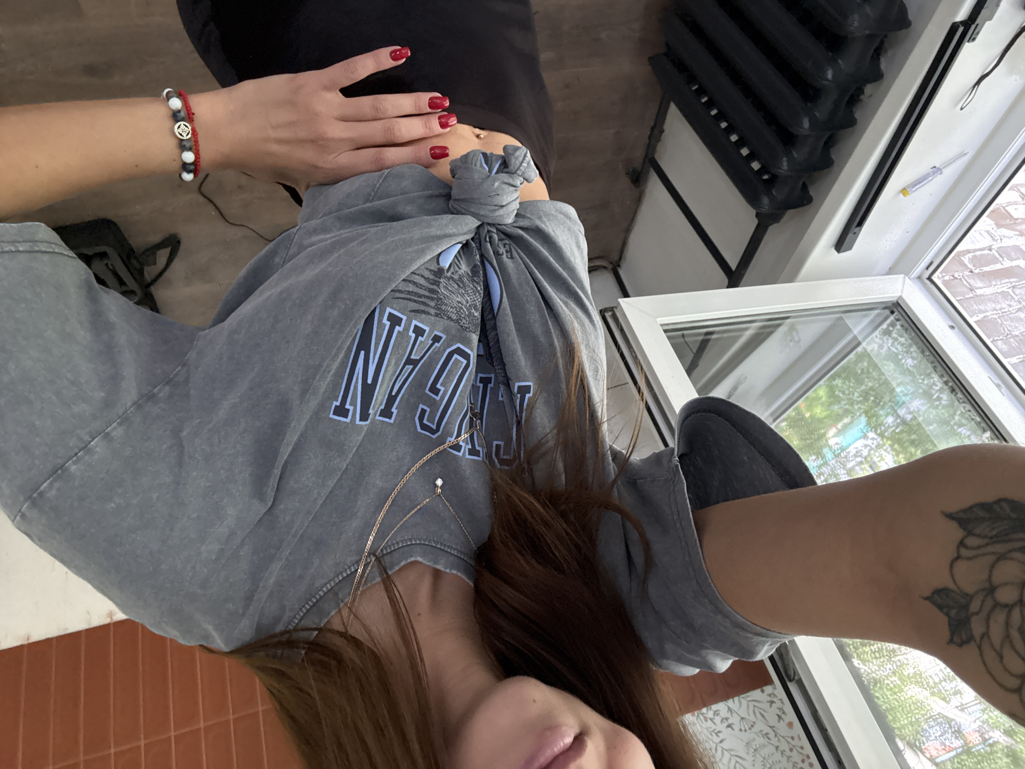 This media file is a horizontal, medium shot of a person, likely a woman, in an indoor setting with a window. The subject is wearing a grey, knot-tied t-shirt over a black top and grey sweatpants. She has red nail polish and a delicate necklace with a small diamond. On her left wrist, she wears two bracelets: one with multiple beads (white, grey, black) and a charm, and another red string bracelet. She has a tattoo of a rose on her right forearm.
The background shows a tiled floor, a black radiator, and a window with a view of greenery and brickwork, suggesting it might be daytime and fair weather. The scene appears to be a room, possibly a home gym or a casual living space. There is text on the t-shirt that appears to say "NY" in large blue letters, possibly part of a larger logo. The overall mood is casual and relaxed. The subject's face is partially visible, and her expression is neutral. The media file is set in Chaikovsky, Russia.