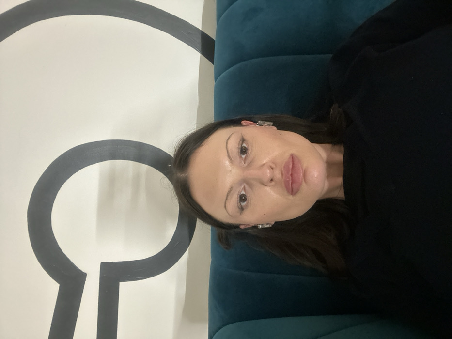 A female individual is seated and photographed indoors on a teal, ribbed upholstered chair. She has long, dark brown hair, dark eyes, and full lips, wearing silver rectangular earrings and a dark, long-sleeved top. Her expression is neutral as she looks directly at the camera. The background is a white wall adorned with a large, dark grey graphic design resembling a broken circle with two linear extensions. This graphic motif is consistent with interior design elements found within The Gherkin building (30 St Mary Axe) in London, United Kingdom.