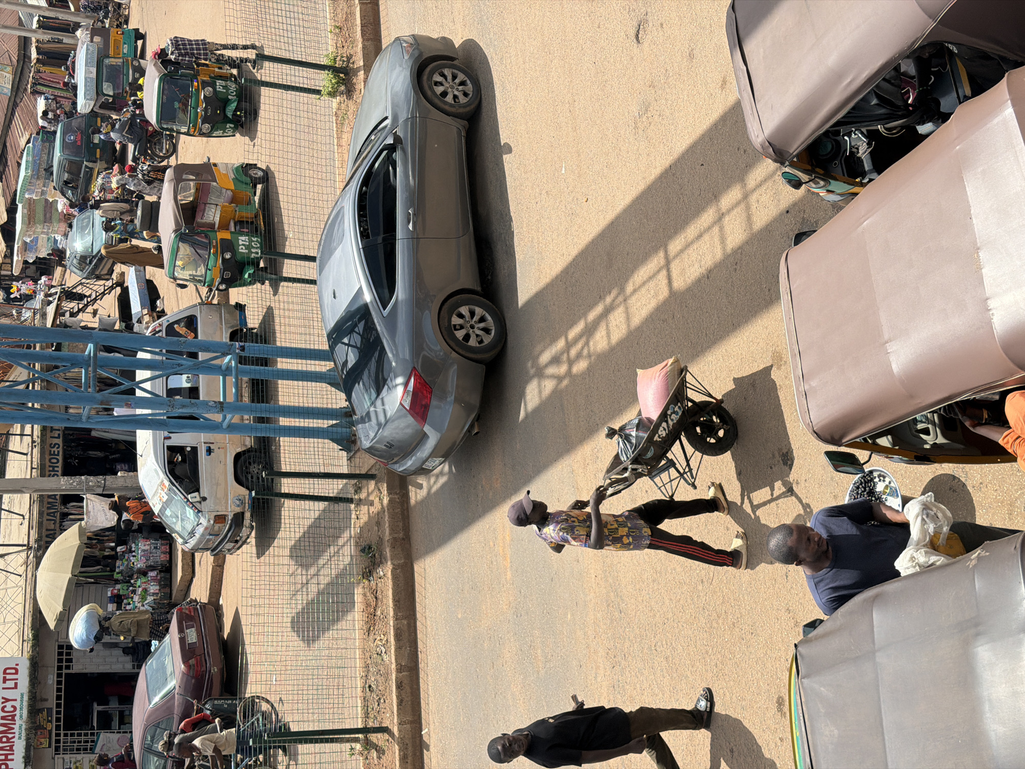 The image depicts a busy street scene in Bukuru, Nigeria, likely during the day due to the strong shadows cast by the sun. The setting is an urban street lined with shops and stalls.
Several vehicles are present, including a grey sedan parked on the road, a white utility van, and multiple three-wheeled auto-rickshaws, commonly known as "keke napeps." Some of these keke napeps appear to be carrying passengers or goods, with colorful awnings providing shade. A handcart loaded with items, possibly for sale or transport, is also visible.
People are engaged in various activities. Some are walking or standing on the street, while others appear to be interacting with the vehicles or stalls. One person is pushing the handcart. Another man is standing next to a keke napep, looking down at some items in a bowl. A man in a black t-shirt is walking away from the camera. Two individuals are seen near a shopfront, with one man in a patterned shirt and cap looking towards the handcart. The presence of awnings and open stalls suggests a market or commercial area.
Visible text includes "PHARMACY LTD." on a sign with "BRUKURU" below it, and potentially a phone number. Another sign reads "MALJAMA SHOES LT".
The overall impression is one of a lively, bustling day in a Nigerian town, with everyday commerce and transportation taking place.