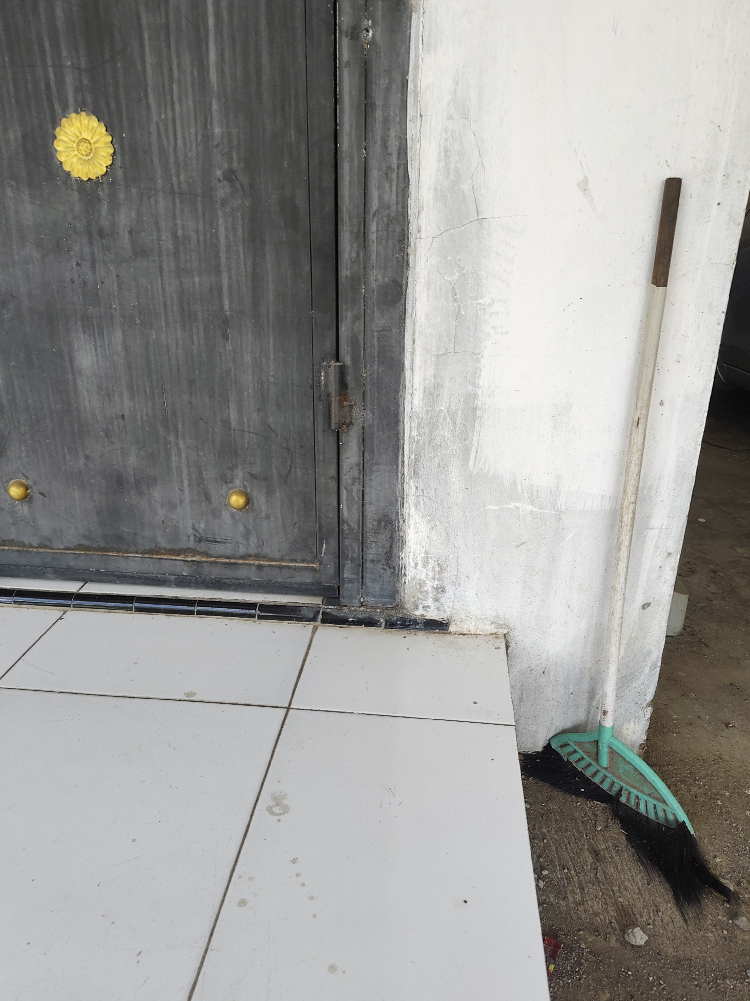 A broom with a teal plastic head and black bristles leans against a cracked, off-white wall next to a weathered gray metal door. The door has two circular brass-colored knobs and a decorative yellow flower emblem on the left side. A tiled floor with light gray squares is visible in the foreground, meeting the base of the wall and door. The ground outside the tiled area is rough and appears to be dirt and gravel. The scene is likely located outdoors, possibly a doorway or entrance in Bangsalsari, Indonesia, given the context.