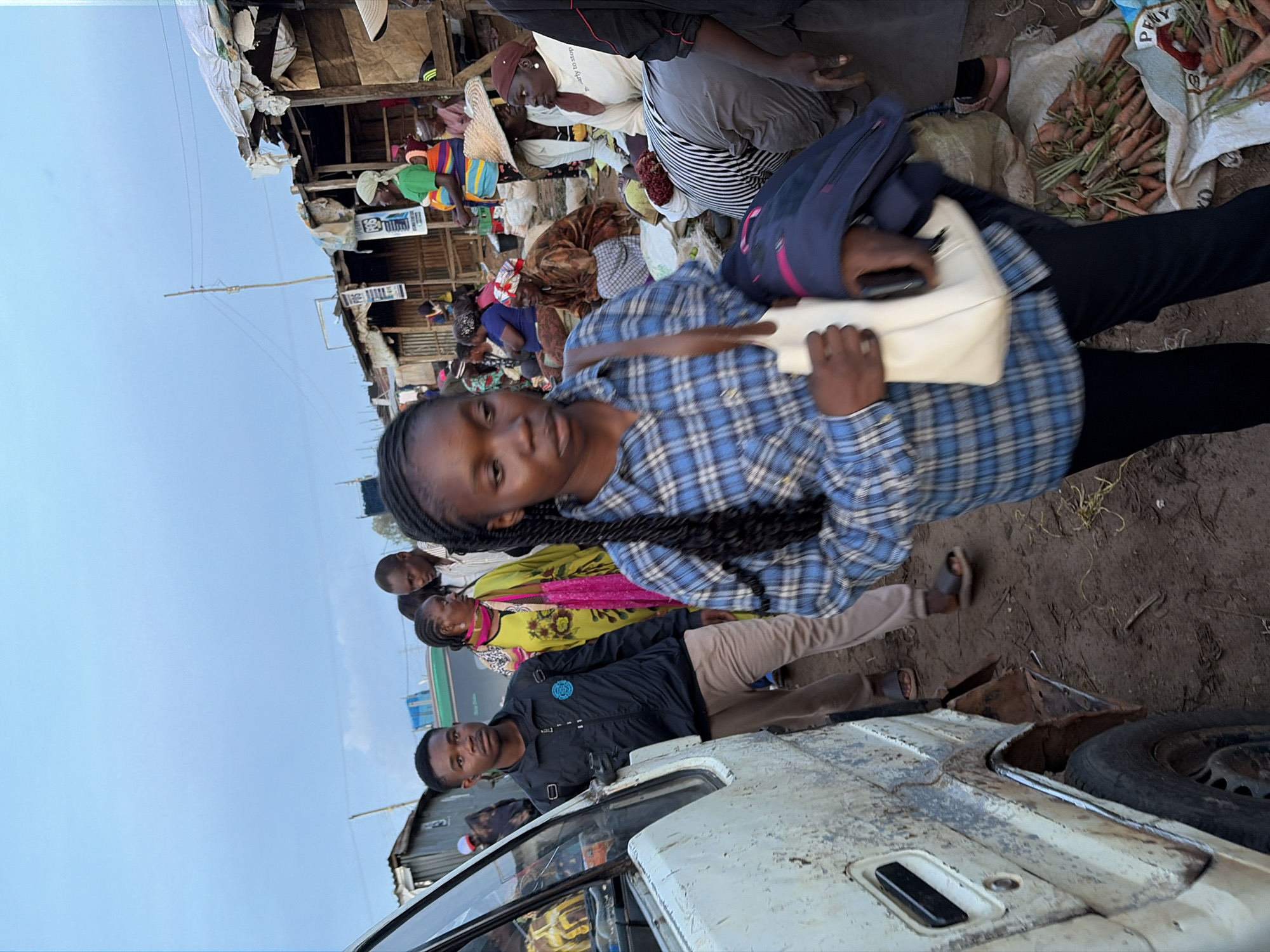 In Gura Top, Nigeria, a bustling open-air market scene unfolds under an overcast sky. A young woman in a blue plaid shirt and black pants, carrying a white handbag and a dark jacket, walks towards the camera. To her left, a man in a black jacket and light brown pants stands near a white van. Other individuals, including women in vibrant clothing and some wearing headscarves, are seen in the background, engaged in market activities. Stalls constructed from wood and corrugated metal line the area, with produce like carrots displayed on sacks. The ground is a mix of dirt and straw.