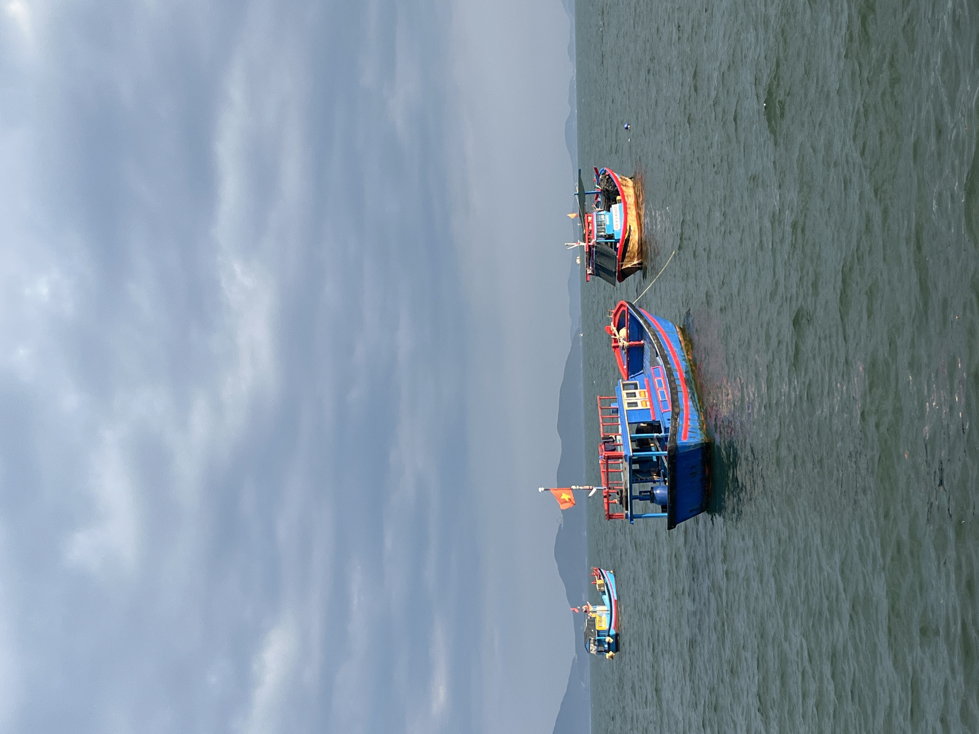 The image depicts a coastal scene in Bac Nha Trang Ward, Vietnam, featuring three brightly colored fishing boats on dark, rippling water under an overcast sky. In the foreground, slightly left of center, a blue fishing boat with a red stripe and yellow accents is moored. It displays a prominent Vietnamese flag (red with a yellow star) on a mast at its stern. Further back on the left, a smaller blue boat with red and yellow details is visible. To the right, another boat, primarily red and yellow-brown with a black canopy structure, is also moored, with a blue buoy floating near its stern. In the distant background, a range of dark, hazy mountains extends across the horizon. The sky above is entirely covered with thick, grey clouds. No individuals are visible on or around the boats.