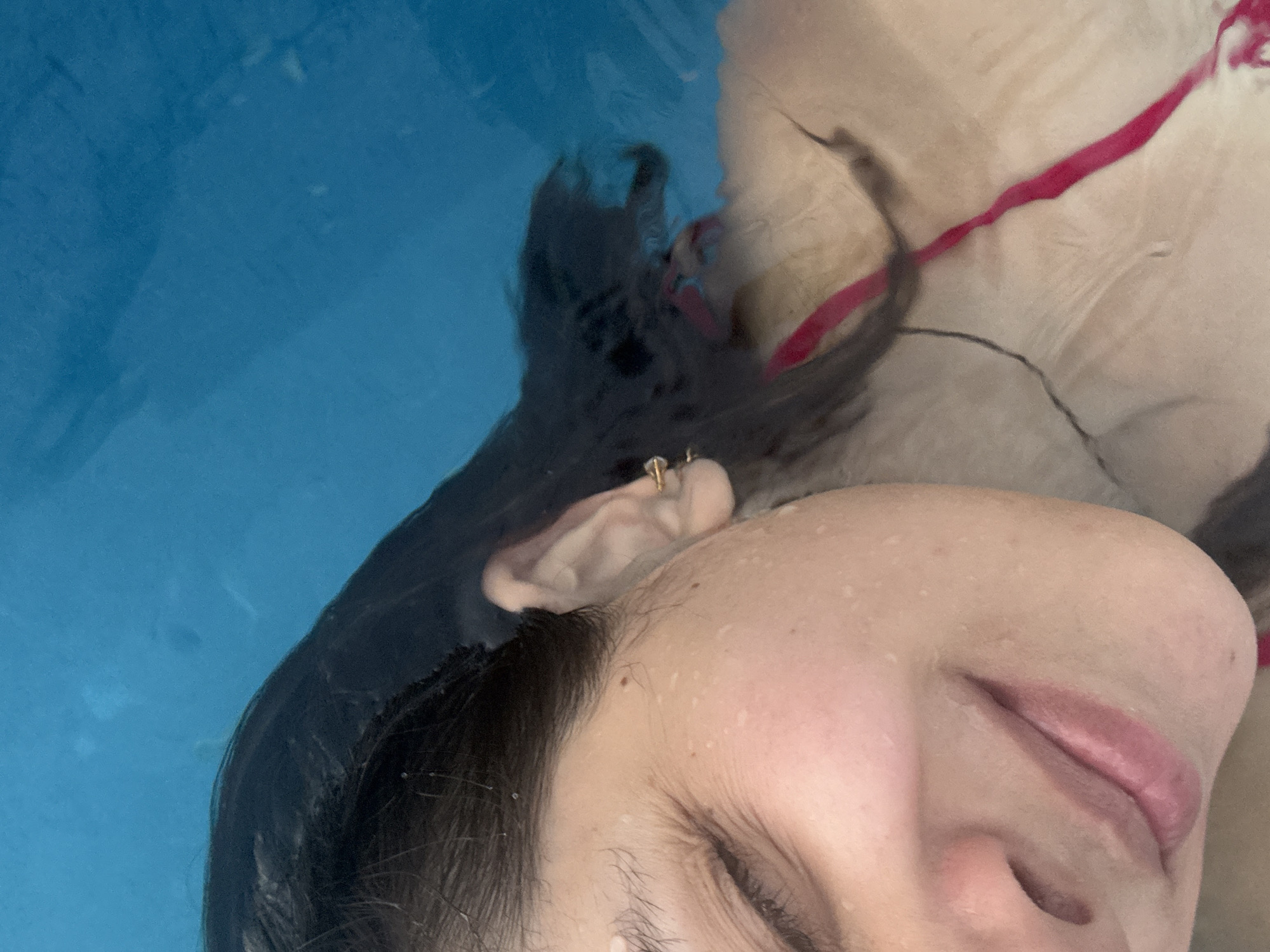 A close-up, slightly rotated image shows a woman partially submerged in a pool of water. Her face, wet with water droplets, occupies the lower right of the frame, with her eyes closed and lips pursed. Part of her dark hair is visible around her face and ear, which is adorned with a gold earring. A red swimsuit strap is partially visible above her shoulder. The background consists of the blue-colored pool water. It is not possible to determine the location of the scene in Veracruz City, Mexico from this image alone.