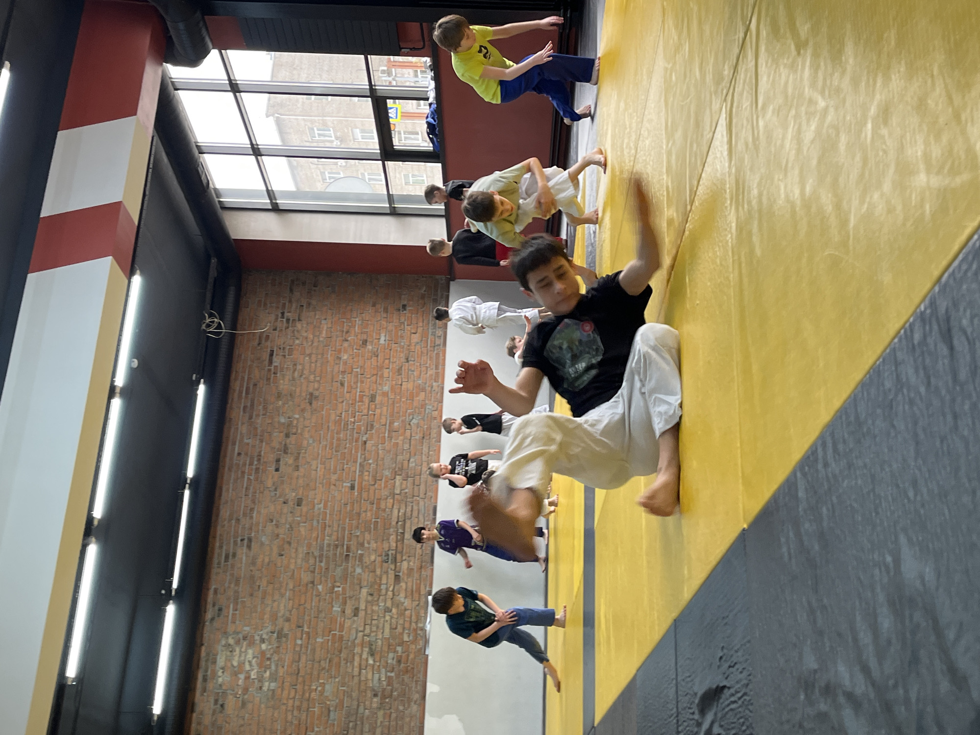 A group of children are practicing martial arts on a yellow and gray mat in a room with a brick wall and a large window. The room is brightly lit by fluorescent lights. The image was taken in the city of Krasnoyarsk, Russia. One child in the foreground is wearing a black shirt and white pants, and is in the middle of a move. Other children are standing and moving in the background. Some of the children are wearing martial arts uniforms.