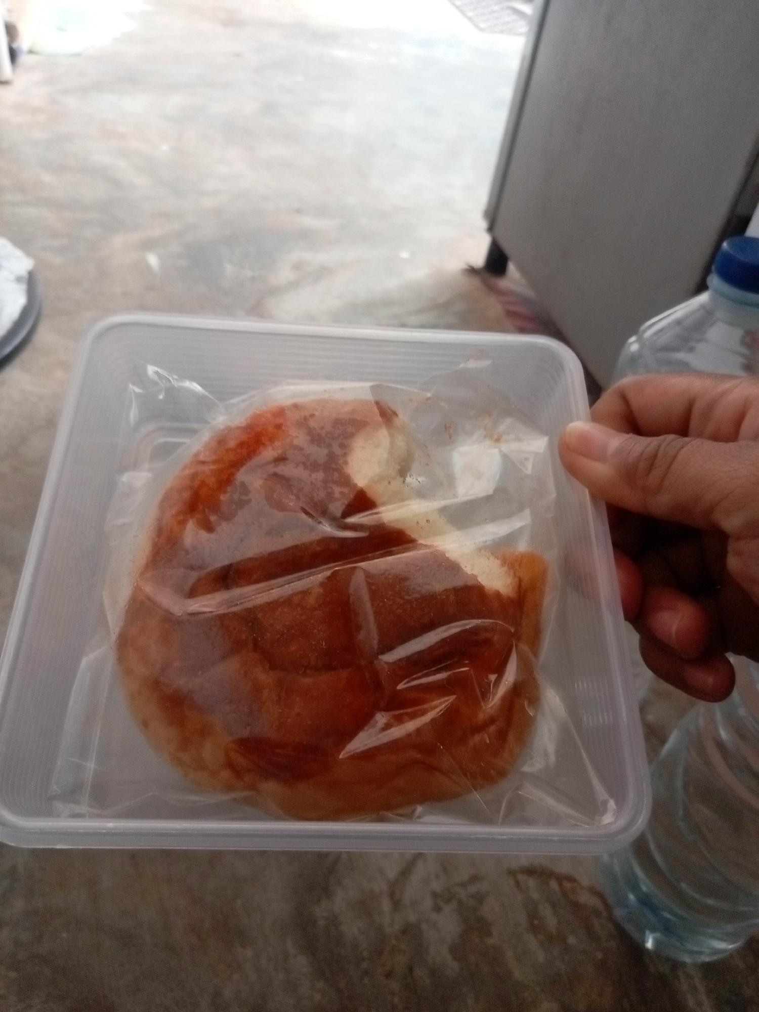 A close-up, overhead shot shows a person's hand holding the corner of a clear plastic food container. Inside the container is a single, round, light brown bread roll, partially wrapped in clear plastic. The bread has a slightly glossy, reddish-brown crust with some lighter sections, suggesting it may have been glazed or toasted. The container itself is rectangular with rounded corners and appears to be made of translucent plastic.
In the background, a portion of a white and gray refrigerator is visible on the right side of the frame. To the right of the food container, a clear plastic bottle of water with a blue cap is partially in view. Another water bottle is visible at the bottom right, also partially filled with water. The ground beneath the objects is a mottled gray surface, possibly concrete or pavement. The scene appears to be outdoors or in a covered outdoor area. The lighting is natural and diffuse. The overall impression is one of casual dining or food preparation.
This image was taken in Batam, Indonesia.