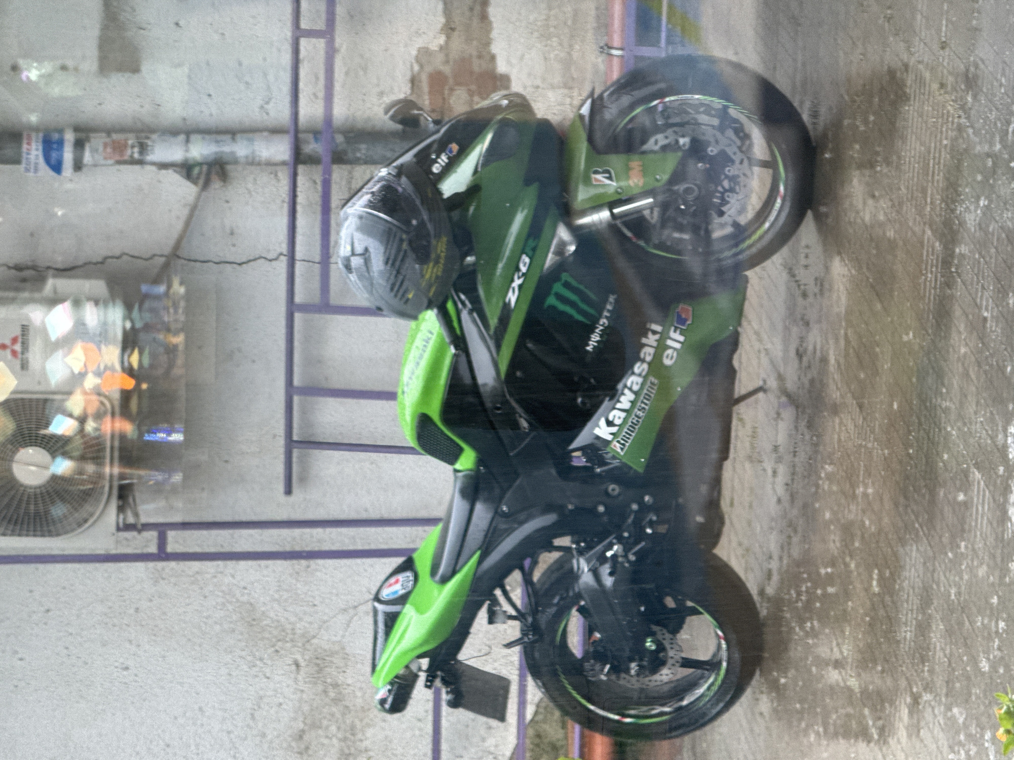 The image depicts a green and black Kawasaki ZX-6R motorcycle parked on a paved surface that appears wet, likely due to rain. A dark-colored helmet rests on the motorcycle. The background shows a building facade with an air conditioning unit and a window or reflective surface through which the motorcycle is being viewed. The scene is likely in an urban setting, potentially Plovdiv, Bulgaria, but there are no immediate visual cues confirming the specific city or country. The weather seems overcast or rainy. There are no visible people.