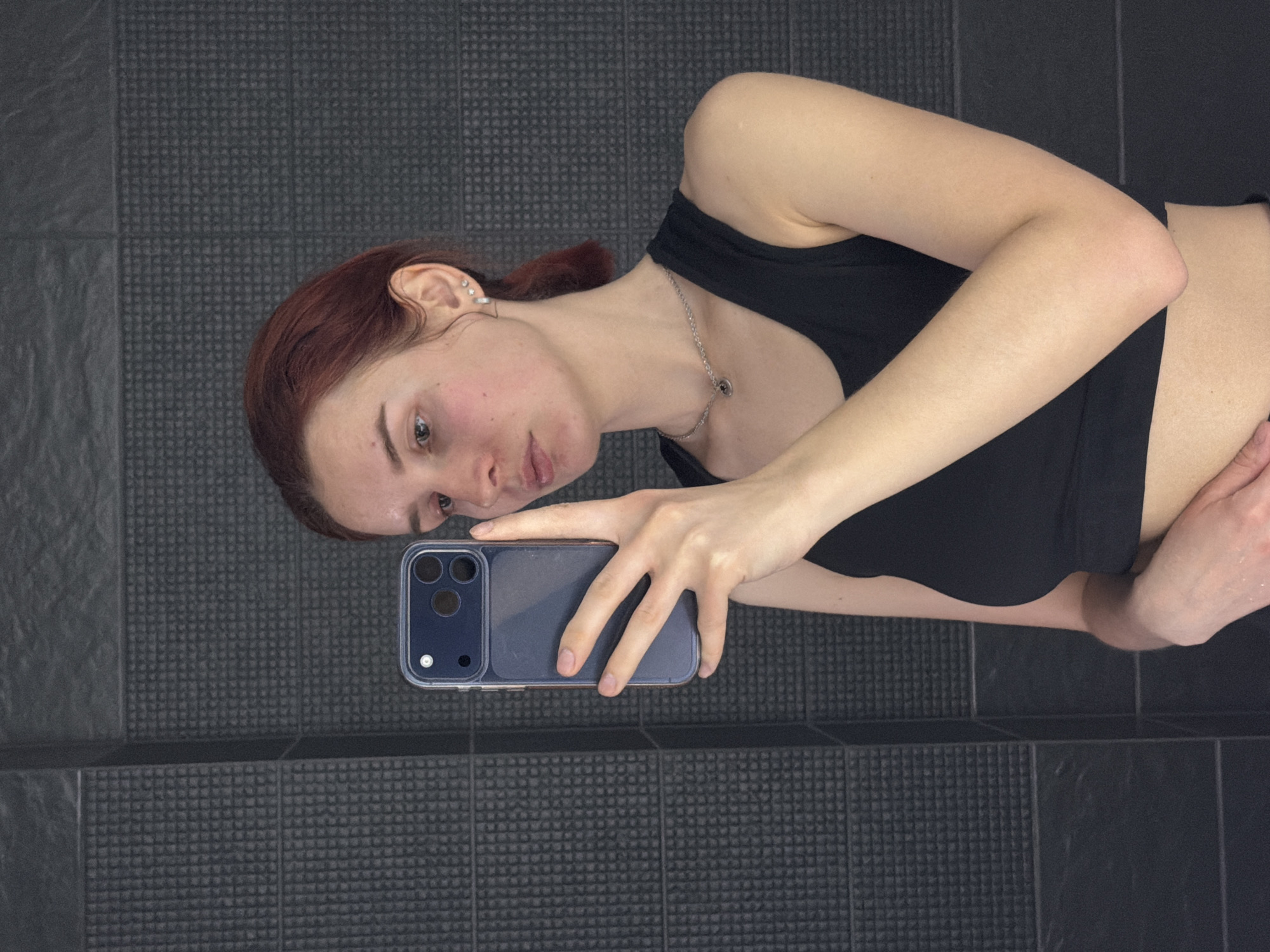 A woman is taking a selfie in what appears to be a bathroom. She is wearing a black sports bra and a silver necklace. Her hair is reddish-brown and pulled back. She is holding a dark-colored smartphone with a clear case in her right hand. The background consists of dark gray tiled walls and floor. The location is unidentified; there's no information to indicate that the photo was taken in Chaikovsky, Russia.