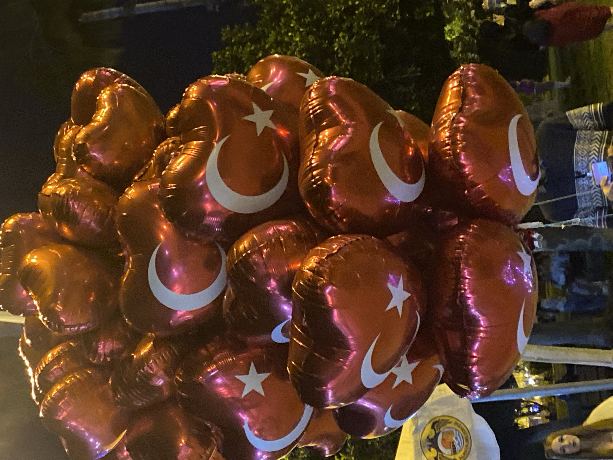 A collection of red, heart-shaped balloons adorned with the white crescent moon and star of the Turkish flag are bunched together. These balloons appear to be in Alanya, Turkey, based on the flag imagery. The background shows a dimly lit outdoor setting at night, with blurred figures of people, some foliage, and urban elements like railing or lighting poles visible. The atmosphere implies a celebration or public event.
