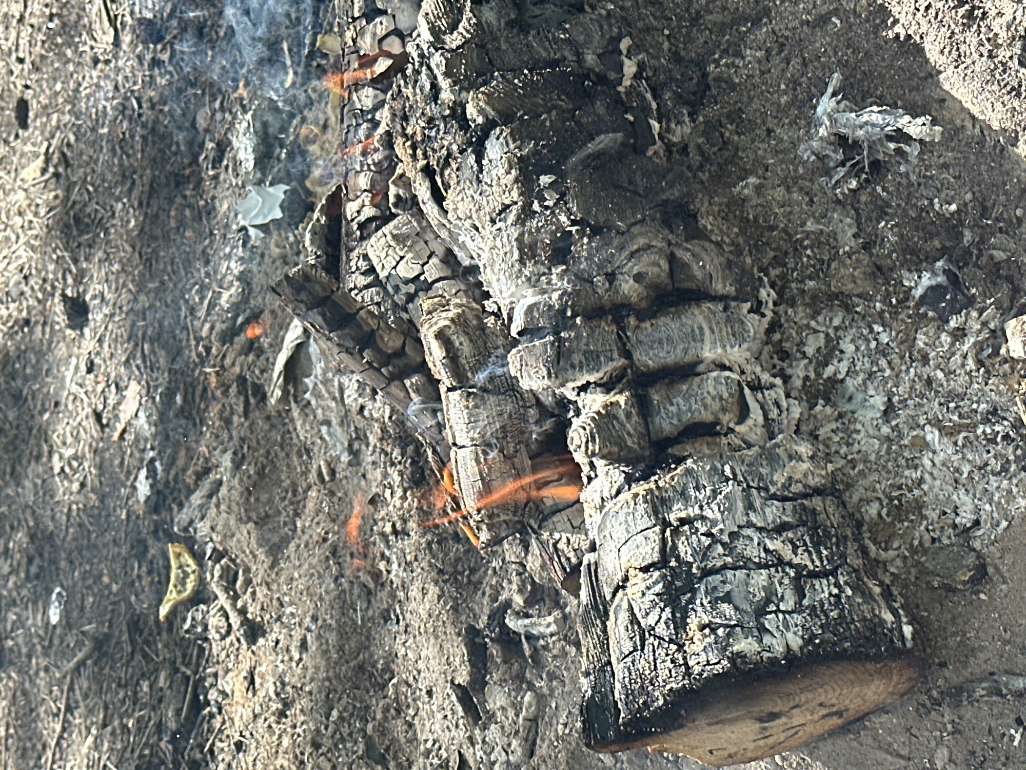 The image depicts a close-up view of burning wood and ash. The wood is charred and cracked, with visible flames and smoke emanating from within. The surrounding ground is covered in ash, debris, and sand. The location cannot be determined from the visual information. There is no evidence of people, interactions, or the location in the city of Юзуфоўскі сельскі Савет, Belarus.