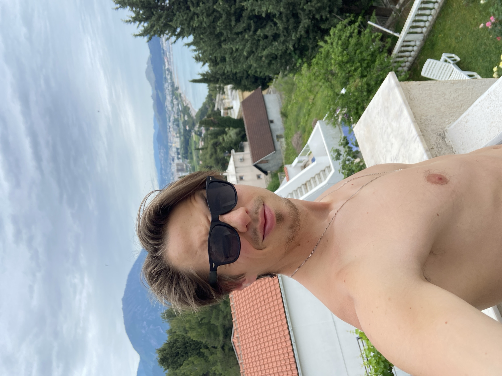 A selfie-style photograph taken outdoors in Šušanj, Montenegro. The image is slightly angled, presenting a man wearing black sunglasses with a fair complexion. He is shirtless, revealing a portion of his upper torso, shoulder, and chest. A thin, silver necklace is visible around his neck.
In the background is a coastal cityscape with buildings and greenery. Further in the background, mountains are partially visible. The sky is overcast with gray clouds. The location is suggestive of a higher elevation point overlooking the city and coast, capturing residential buildings, vegetation, and the distant sea.