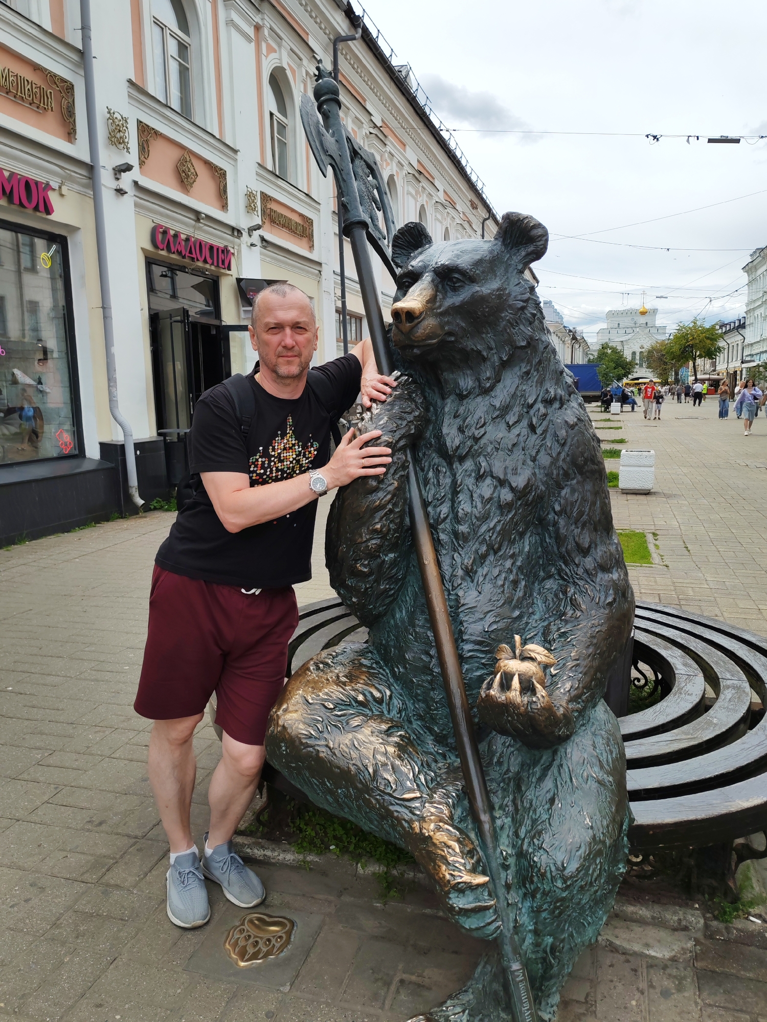 The image shows a man posing with a bronze statue of a bear in Yaroslavl, Russia. The man, with a bald head and a beard, is wearing a black t-shirt with a colorful geometric print, maroon shorts, and grey sneakers with white socks. He is leaning on the statue of the bear, which is seated and holding a staff with an axe-like head and an apple in its paws. A bronze bear paw print is visible on the pavement in front of the statue.
The statue is located on a paved street lined with buildings. The buildings have ornate architecture with arched windows and decorative elements. Signs on the buildings are in Cyrillic script. One sign visible says "СЛАДОСТИ" which translates to "Sweets". In the background, more buildings and people walking can be seen, suggesting this is a public area, likely a pedestrian street. The sky is overcast, indicating a cloudy day. The time of day appears to be daytime. The overall mood is casual and touristy, with the man posing for a photo with a local landmark.