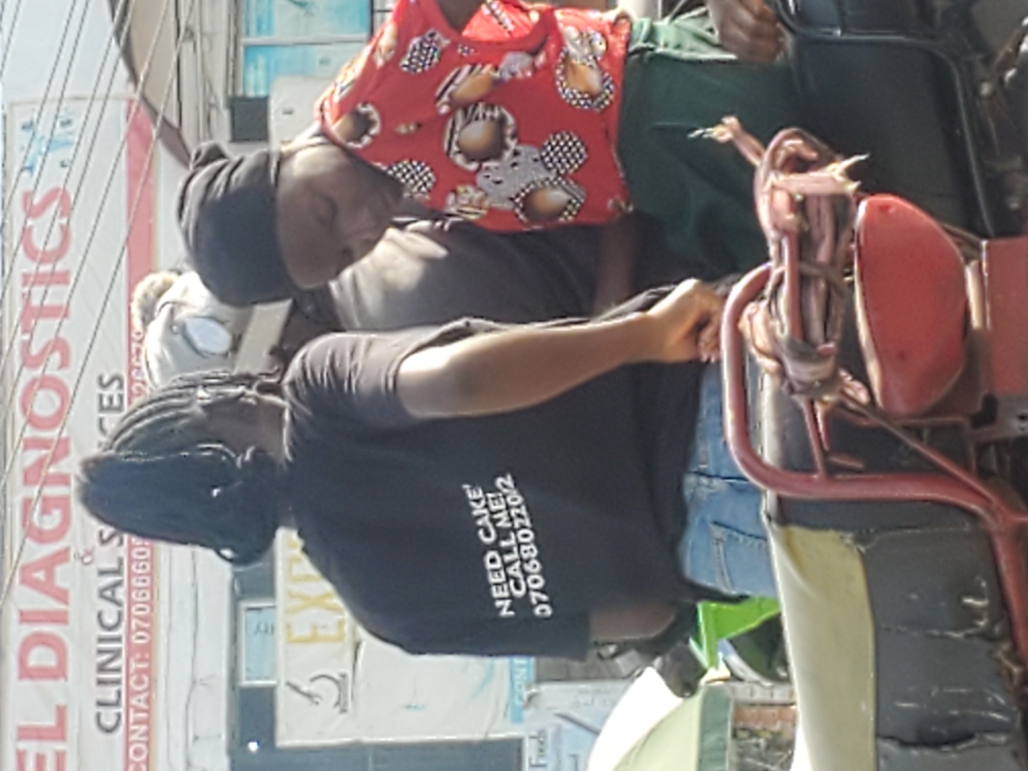The image shows three people on a motorcycle. The person in the front is wearing a black t-shirt with the text "NEED CAKE! CALL ME! 0706802202/2" printed on the back. The person in the middle is wearing a black headscarf and a red and white patterned shirt, and is looking down. The third person is wearing a grey shirt and a white helmet. They are in a city environment, with buildings and signs visible in the background, including a sign for "EL DIAGNOSTICS". The motorcycle has a red frame and a light-colored seat. It is daytime, and the lighting suggests an outdoor setting. The location is identified as Gwong, Nigeria.