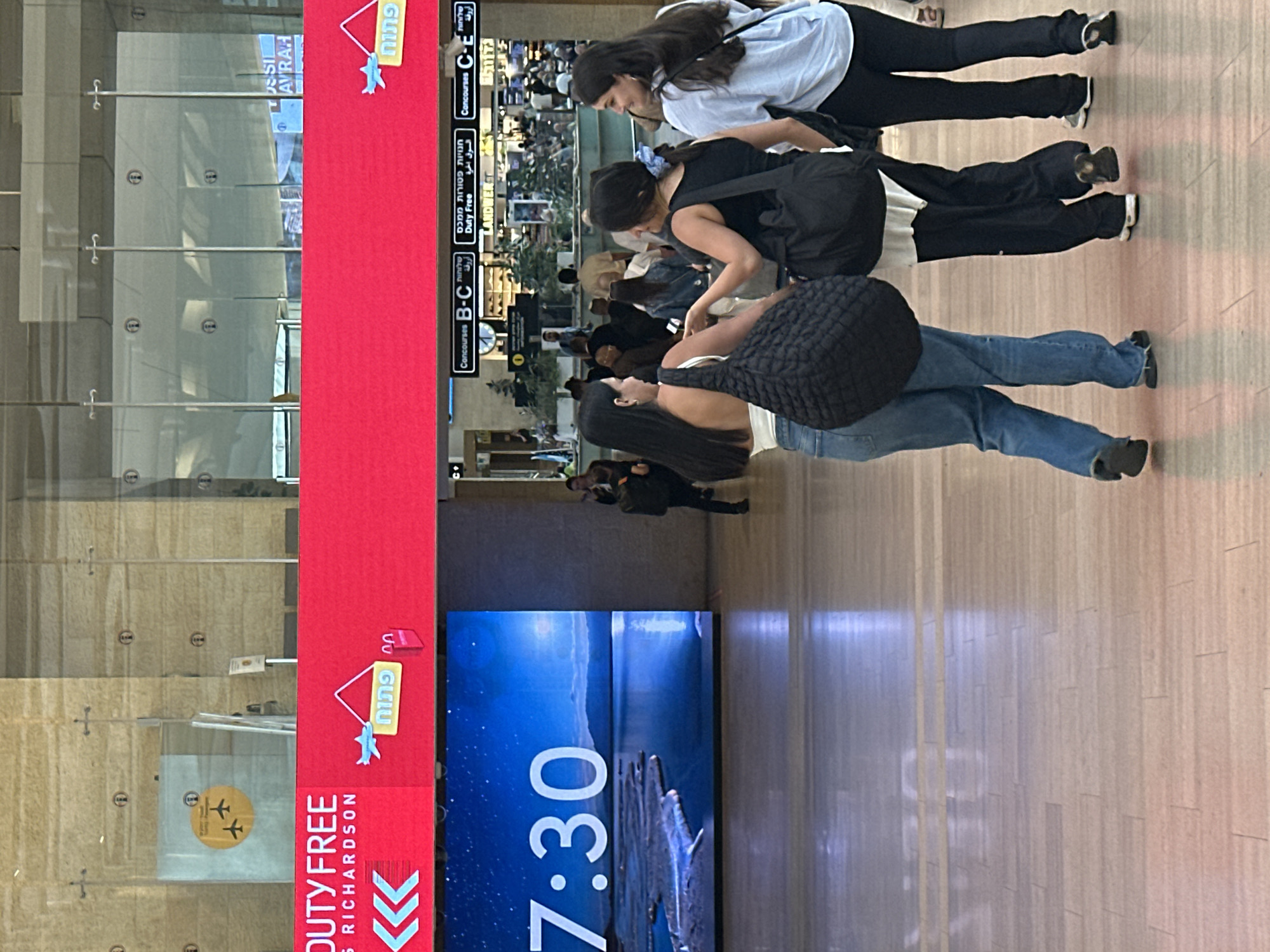 The image captures a scene in Terminal 1 of Ben Gurion Airport, located in Tzafria, Israel. A large, bright red digital display board dominates the upper portion of the frame, featuring the text "DUTY FREE" and a shopping bag icon. Below this, a digital clock displays "7:30" against a serene blue background of water and mountains. In the foreground, three young women are walking away from the camera. The woman on the left wears a white top and blue jeans, carrying a large black quilted bag. The middle woman has dark hair and wears a black top and dark pants, also with a black bag. The woman on the right is in a light blue shirt and black pants, with a black bag slung over her shoulder. They are walking on a polished wooden floor in what appears to be a modern airport terminal. In the background, airport signage indicates concourses and a "Duty Free" shop. Other travelers are visible in the distance, adding to the bustling airport atmosphere.