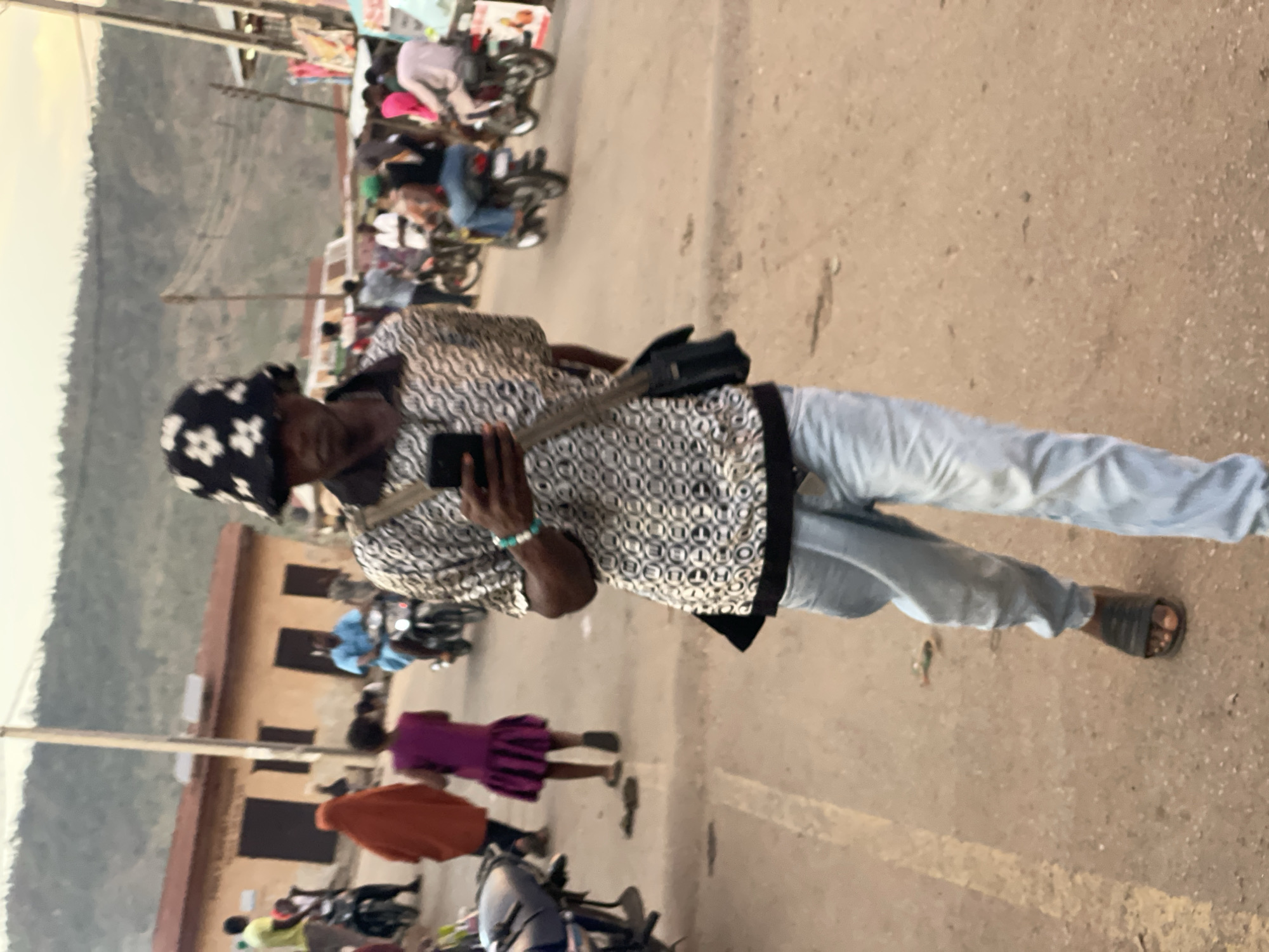 A man stands in the foreground, holding a smartphone and wearing a patterned shirt, light blue jeans, a head covering, and sandals. A black bag hangs across his torso. In the background, a street scene unfolds with several people and motorcycles. Buildings line the street. The scene is set in Lokoja, Nigeria. The man is the primary subject of the image. Other figures are more distant and less detailed.