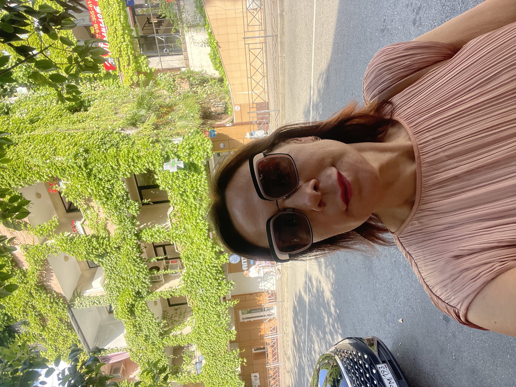 The image is a selfie taken by a woman in front of a building covered in green ivy in Sochi, Russia. The woman is wearing large sunglasses and a pink pleated top. Behind her, the building has balconies and windows, with air conditioning units visible on some of the walls. A black car with a license plate that reads "PM 193 RUS" is parked to her left. The street is paved, and there is a sidewalk with a metal fence in front of the building. In the background, there are shops and signs, including one with Russian text and a phone number. The overall scene suggests a sunny day in an urban environment.