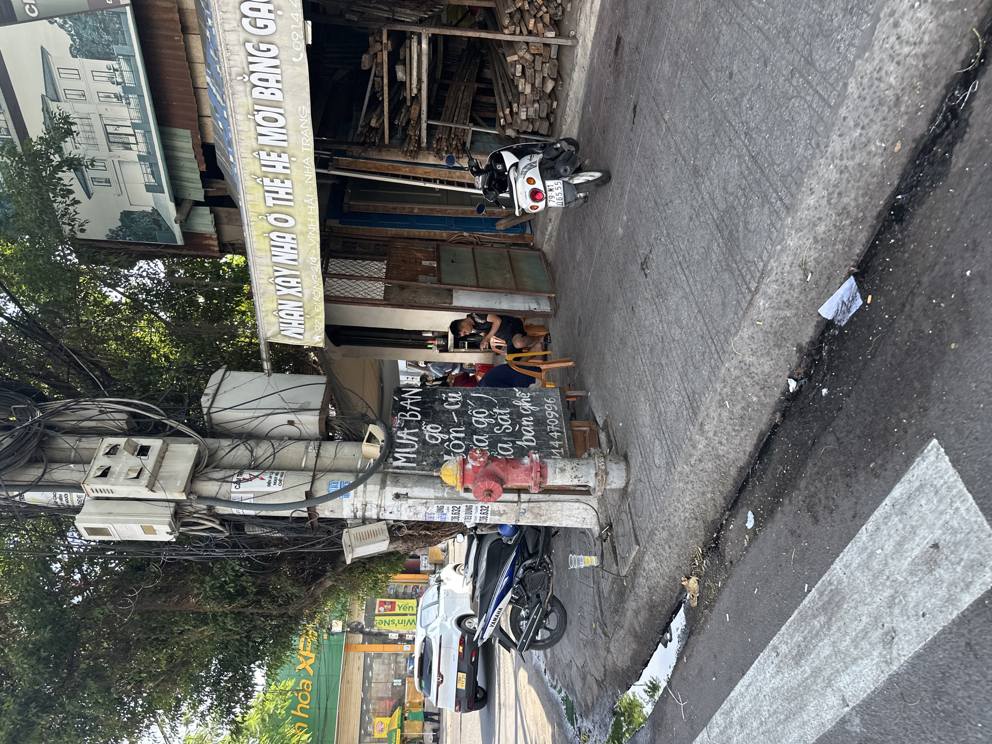 The image depicts a street scene in Bac Nha Trang Ward, Vietnam, characterized by a busy sidewalk and road. A prominent utility pole covered in a dense network of wires stands on the sidewalk, adorned with several electrical boxes and conduits. Nearby, a red and yellow fire hydrant is visible. A black and blue Yamaha scooter is parked on the sidewalk, partially obscuring a small table with a plastic cup and a clear liquid.
A chalkboard sign on the sidewalk displays Vietnamese text, seemingly advertising items for sale. A young man sits on an orange chair next to the chalkboard, looking at his phone. Further down the sidewalk, another man is seated, also looking at his phone. In the background, a white SUV is parked on the road, and beyond that, a green building with signage is partially visible. A white scooter is parked on the sidewalk in front of a building with stacked lumber visible through an open doorway. A banner overhead reads "NHAN XAY NHA O THE HE MOI BANG GẠO," translating to "Building houses for the new generation with rice."
The environment appears somewhat cluttered, with wires hanging from the utility pole, some debris on the sidewalk and road, and stacks of building materials. The time of day seems to be daylight, with shadows cast on the ground. A pedestrian crossing is visible in the foreground on the asphalt road.