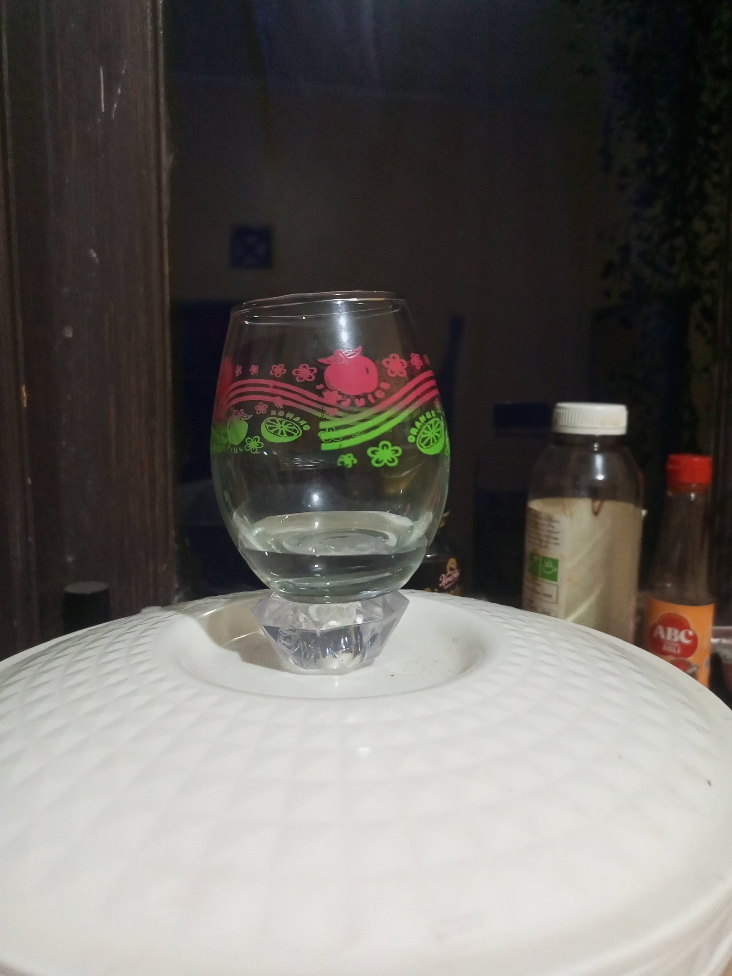 A clear glass tumbler with a pink and green fruit design rests on a diamond-patterned white surface. The tumbler contains a small amount of clear liquid. Behind the tumbler, two bottles are visible: a taller, opaque bottle with a white cap and a smaller, orange bottle with a red cap. The background is dark, with some indistinct shapes suggesting indoor furniture and foliage. The scene is captured from a low angle, emphasizing the tumbler and the white surface it sits on. The location is identified as Cidahu, Indonesia.