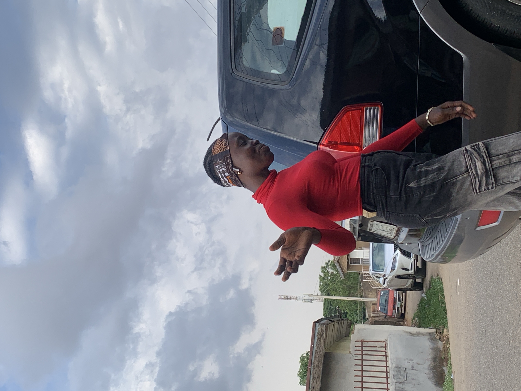 A person in a red turtleneck and grey jeans is positioned at an angle leaning against the rear of a black SUV. The background includes a street with parked cars, buildings, and a cloudy sky. The location is likely in Jos, Nigeria, given the style of architecture and the overall environment. The person is wearing a patterned headband and a bracelet.