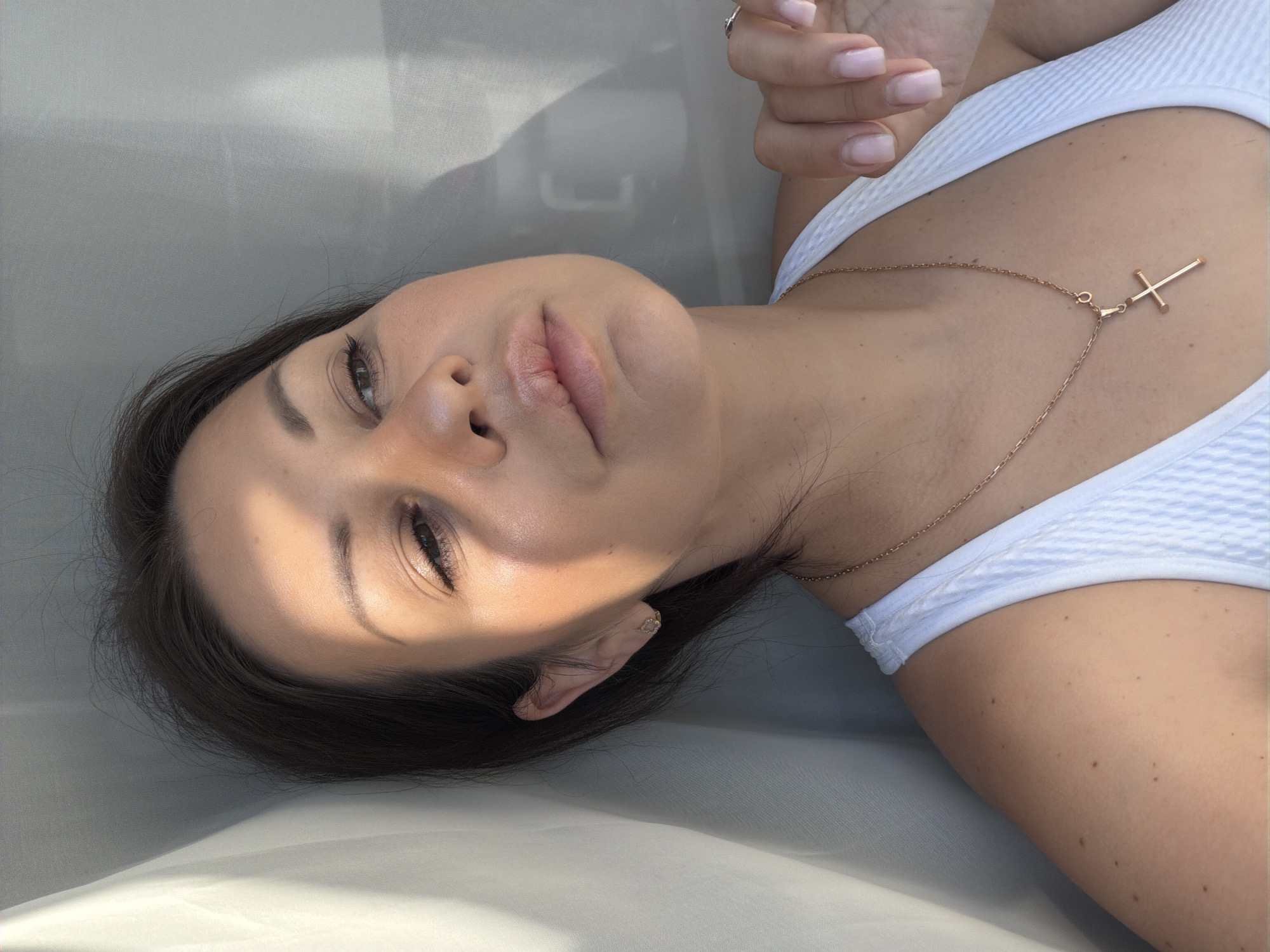 In a close-up portrait, a woman with dark hair and light skin is positioned in what appears to be a sun-drenched indoor setting, possibly a room in Sochi, Russia. Sunlight streams across her face, illuminating the left side of her forehead and cheek, creating a dramatic interplay of light and shadow. Her eyes are partially closed, and her gaze is directed slightly upward and to the right. She is wearing a white tank top and a delicate gold necklace with a cross pendant. Her left hand is visible, resting on her chest, with a ring on her ring finger. The background is a soft, out-of-focus white material, likely a curtain or fabric, which is partially illuminated by the same sunlight. The overall impression is one of quiet reflection and natural light.