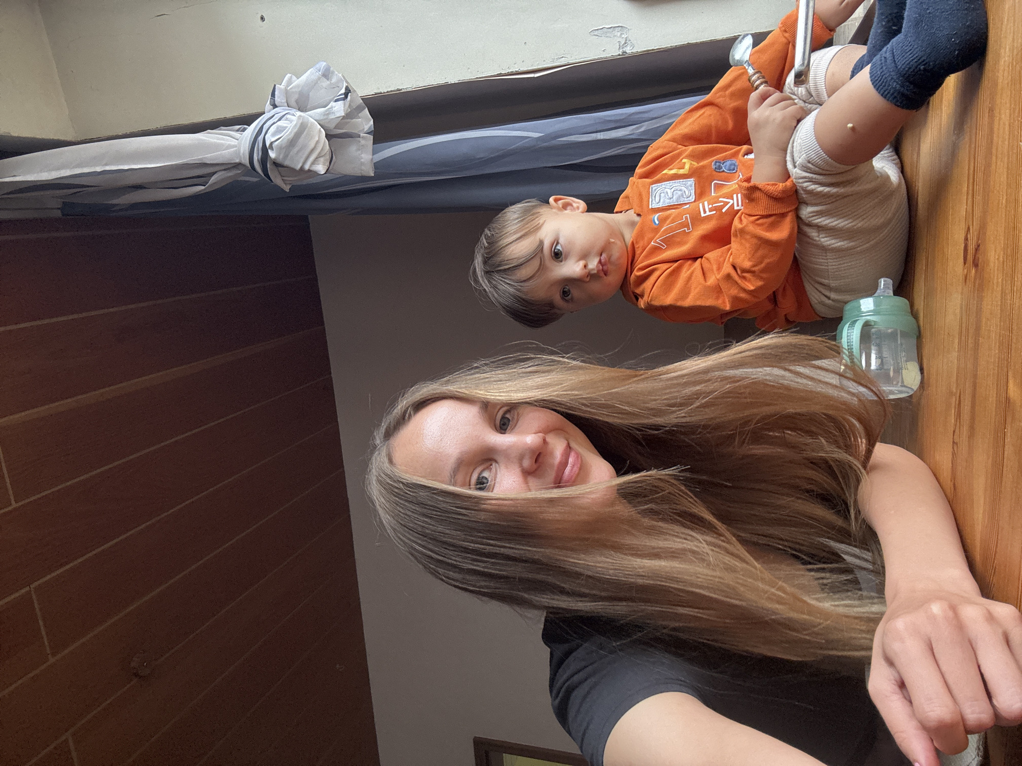 In an indoor setting that appears to be a home in Überherrn, Germany, a woman with long blonde hair and a black t-shirt is positioned in the foreground, resting her arms on a wooden surface. To her right, a young child wearing an orange hooded sweatshirt and light-colored pants is seated, holding a spoon in their right hand. The child has a small white speck on their knee and a drip of liquid on their chin. A baby bottle with a green lid is visible on the wooden surface in front of the child. The background features a wooden slatted ceiling and a window with a striped curtain tied back. The overall lighting suggests it is daytime.
