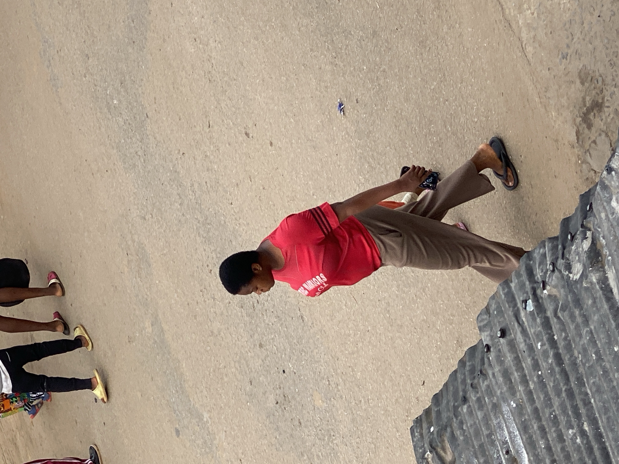 A photograph shows a person wearing a red shirt and brown pants standing on a concrete surface in Fegge, Nigeria. The person is holding a phone in their hand and is looking down. A group of people are to the left, and there is a corrugated metal surface to the right. A small piece of blue trash is on the ground near the standing person.