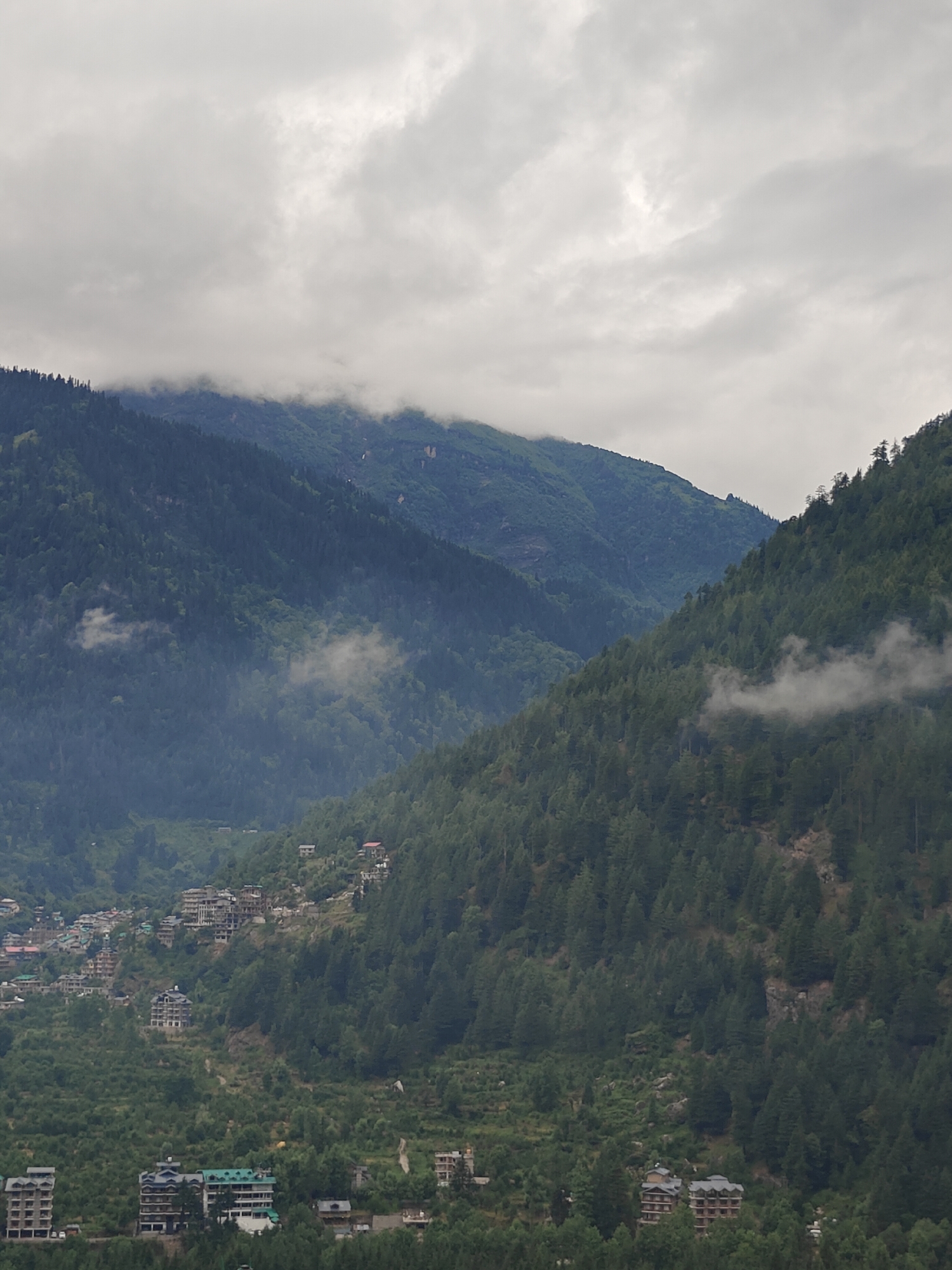 The image depicts a mountainous landscape with a town nestled on the slopes, likely in Vashisht, India. The sky is overcast with thick, gray clouds. Dense green forests cover the steep mountain sides. In the midground and foreground, buildings of various sizes and architectural styles are scattered across the terrain, some appearing to be hotels or residences. Patches of mist or fog are visible, adding to the atmospheric conditions. The overall scene conveys a sense of a rural or semi-urban settlement within a majestic natural environment.