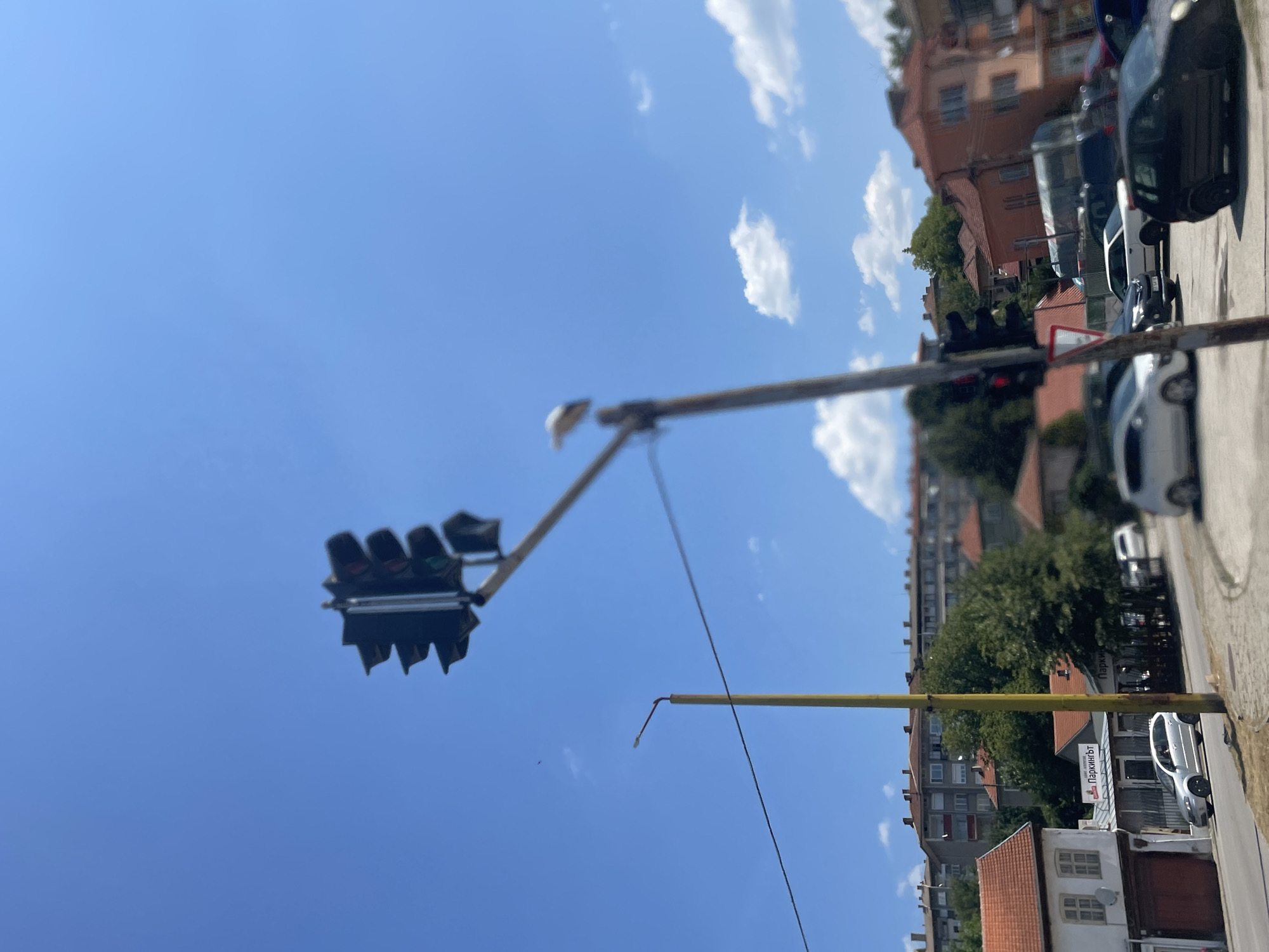 This is a medium shot, outdoor, daytime, eye-level, full-color, slightly out-of-focus photograph of a street scene in Gabrovo, Bulgaria. In the foreground, a traffic light pole with a functional traffic light is visible, with a bird perched on the horizontal arm of the traffic light. Behind the traffic light, a yellow lamppost is also visible. The background features buildings of various architectural styles, including multi-story apartment buildings and smaller residential homes with red tile roofs. There are trees and a bright blue sky with scattered white clouds. Several cars are parked or driving on the street. The overall atmosphere is one of a typical urban environment on a sunny day.
