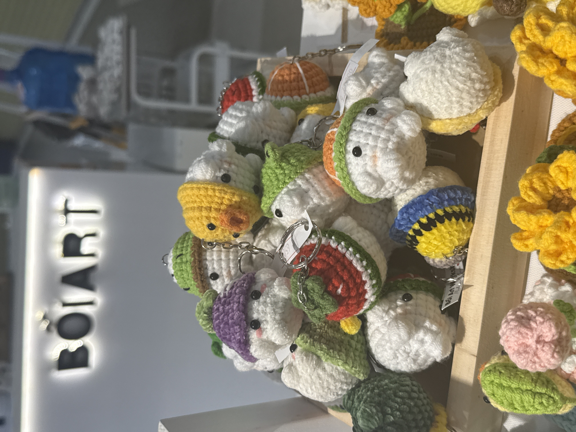 A close-up shot, taken indoors, showcases a collection of handmade, knitted keychains. These keychains are in a variety of shapes and colors, including some designed to look like animals, fruit, and flowers. They are hanging on display, likely for sale. To the left of the keychains is a white wall displaying the vertically oriented, illuminated word "BOIART." The blurred background suggests the location is a shop or market. The shot was taken in Nha Trang Ward, Vietnam.