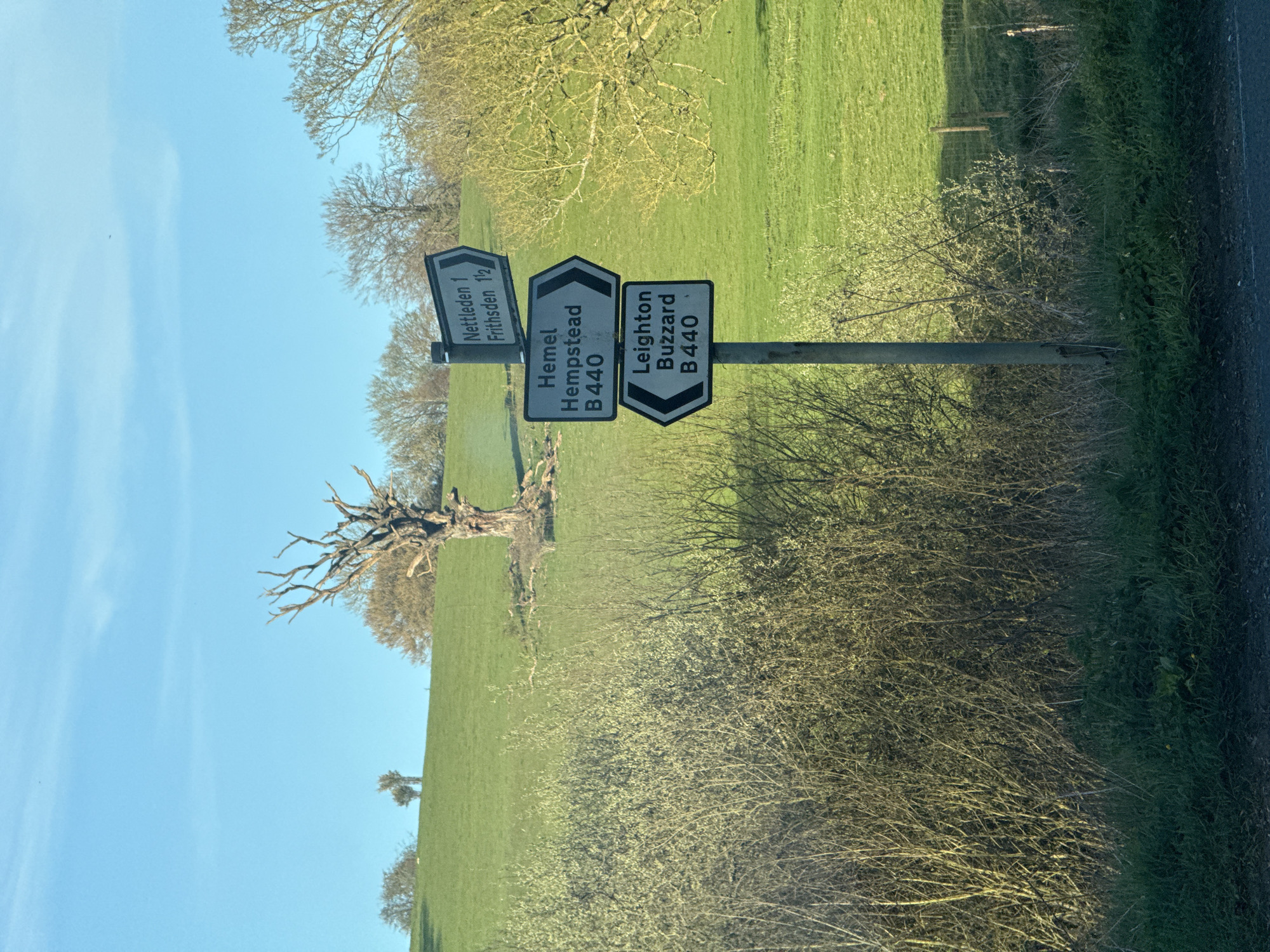 The image shows a rural roadside scene in the Dacorum region of the United Kingdom. A signpost stands prominently, indicating directions to "Nettleden 1 Frithsden 1 1/2," "Hemel Hempstead B440," and "Leighton Buzzard B440." The signpost is positioned beside a road with overgrown vegetation. Beyond the road, a grassy field stretches out with a solitary, bare tree. The sky is blue with slight cloud cover.