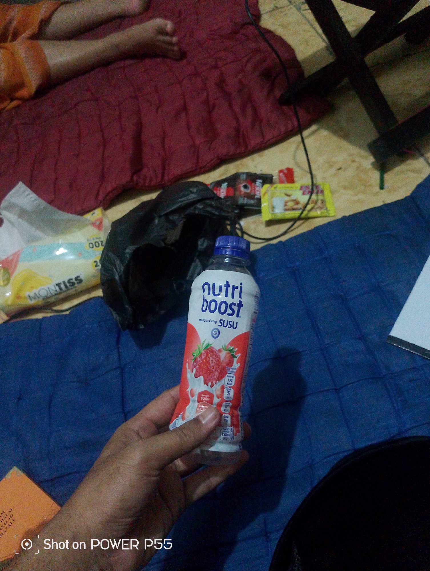 In Lebaksiu, Indonesia, an individual is holding a bottle of NutriBoost strawberry-flavored milk. The bottle is white with red and pink branding, featuring an illustration of strawberries. The background consists of a red quilted mattress, a blue mat, a black plastic bag, and a few other items including a pack of Montiss wet wipes and what appears to be Nescafé sachets. The scene suggests a casual indoor setting, possibly a bedroom or a living area. The lower right corner of the image shows a dark, fuzzy object, possibly part of clothing or furniture, and the timestamp "Shot on POWER P55".