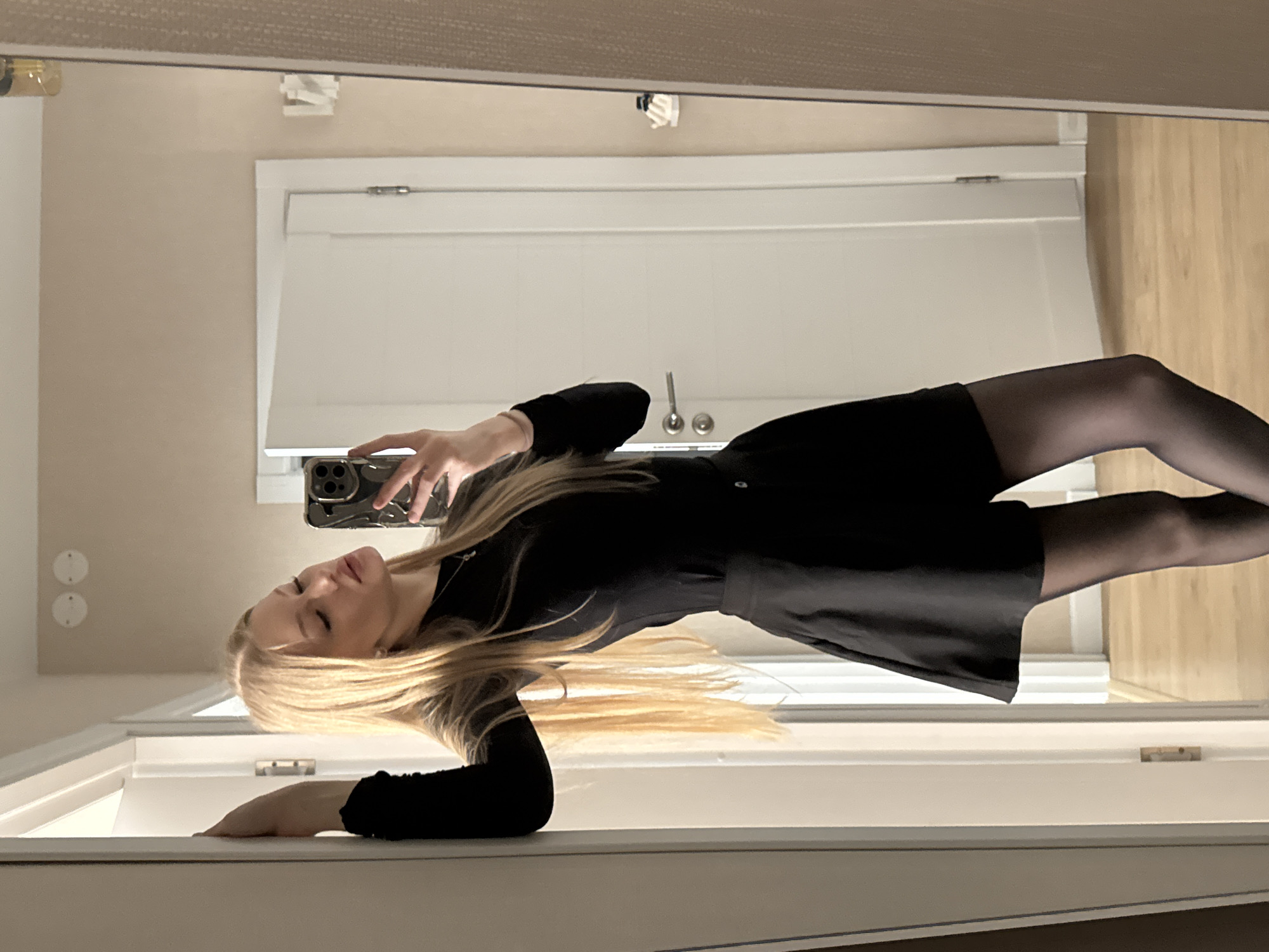 The image captures a blonde female subject taking a mirror selfie within an interior residential or hotel setting, located in Perm, Russia. The subject is positioned with her body slightly angled, wearing a black long-sleeved top, a black mini-skirt, and black sheer tights. Her left hand rests on a white doorframe, while her right hand holds a dark-cased smartphone, likely an iPhone, to photograph her reflection. Her head is tilted upward and to her right, with eyes partially closed and lips slightly parted, indicative of a posed expression.
The background reflected in the mirror displays light-colored wooden flooring and beige painted walls. Two white interior doors are visible; one directly behind the subject is slightly ajar. Two circular ceiling light fixtures are present on the upper portion of the reflected wall. A single light bulb or small pendant fixture and a white wall-mounted object are also visible in the reflection on the right side of the frame.