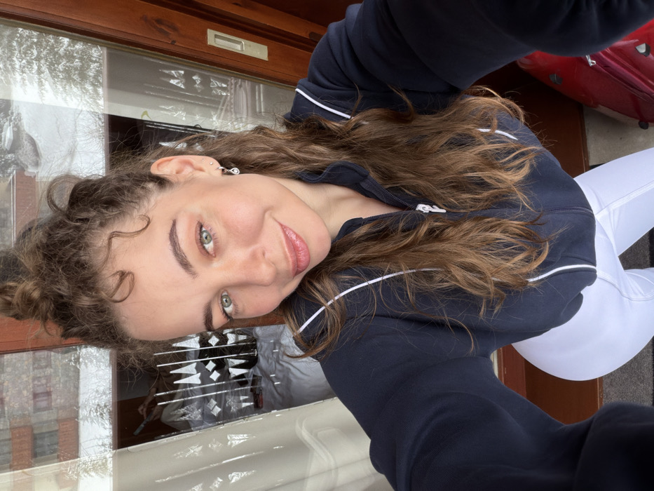 A female individual is depicted in a close-up, high-angle selfie, positioned indoors near a glass door or window. The subject has light skin, green eyes, and light brown wavy hair, partially styled in an updo with loose curls framing her face. She is wearing a dark blue full-zip jacket with white piping and white fitted leggings. A small, teardrop-shaped earring is visible on her left ear. She maintains direct eye contact with the camera and exhibits a slight smile.
The background, visible through the glass on the right and top, presents an exterior winter landscape consistent with the commune of Les Allues, France. This view includes snow-covered evergreen trees and distant buildings with pitched roofs, indicative of a mountain resort environment. The left section of the glass reflects an interior space, revealing a white-sheeted bed, a backgammon board, and several red items, possibly cosmetics. A dark wooden door frame is present on the right, and a partial red object, resembling a piece of luggage, is visible on the floor in the bottom right corner of the frame, adjacent to the female's leg.
