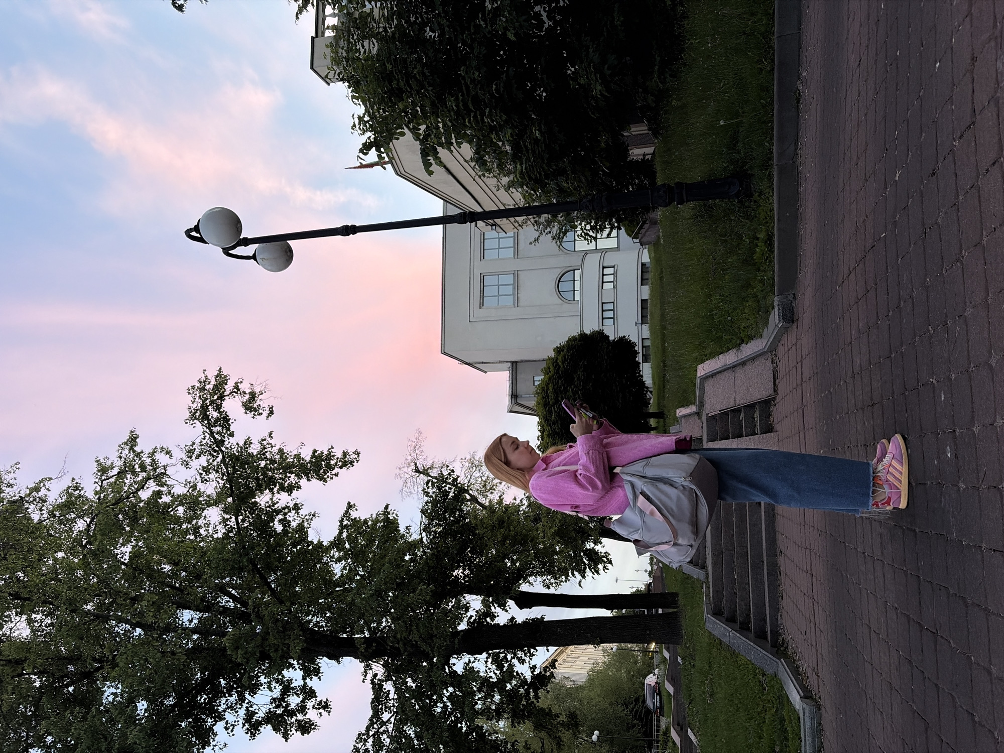 A young woman is captured in an outdoor, vertical-format photograph during what appears to be dusk or evening in Minsk, Belarus. The sky transitions from a light blue at the top to a soft pink horizon, with subtle clouds.
The woman stands on a dark, reddish-brown paved path, made of brick-like blocks, which features several steps leading upwards. She has blonde hair and is wearing a bright pink long-sleeved top, blue wide-leg jeans, and distinctive pink, yellow, and blue sneakers. A light grey shoulder bag is slung over her shoulder. She is actively engaged with her smartphone, holding it with both hands and looking down at its screen.
To her right, a tall black street lamp with two spherical white lights extends into the colorful sky. The path is bordered by lush green grass and large, dark green trees with visible branches and foliage on both sides of the frame. In the mid-ground, behind the trees, a multi-story white building with various rectangular and arched windows is visible, indicating an urban setting. A dark car is partially discernible in the distance further down the path. The scene conveys a calm, serene atmosphere at the end of the day. No discernible text is visible in the image.