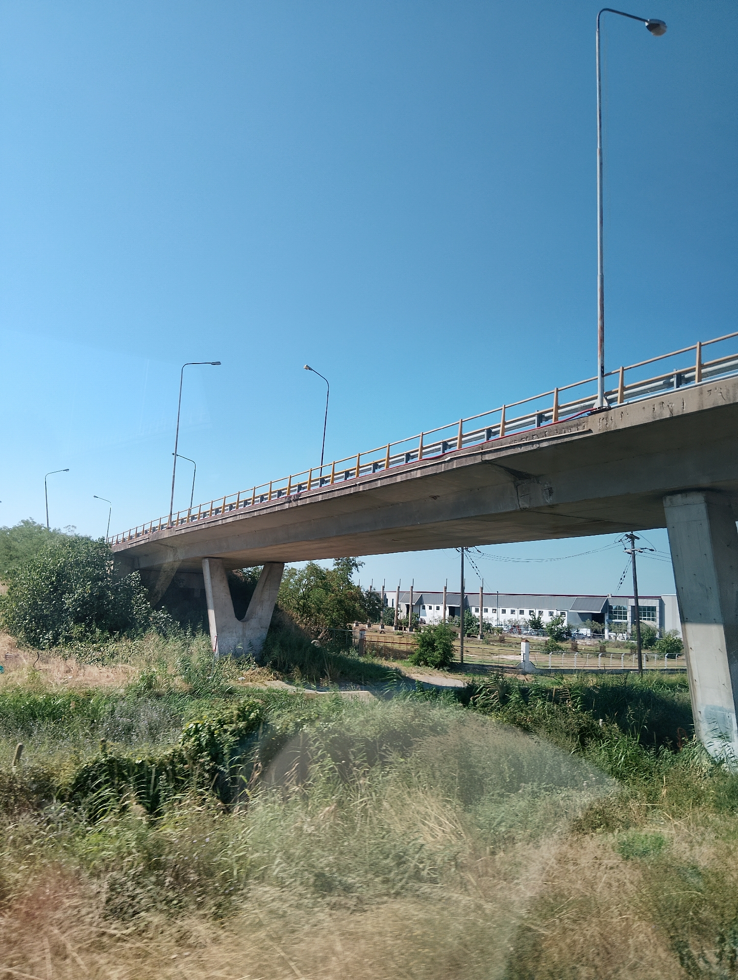 The image captures a concrete overpass spanning a dry, grassy area. The overpass has yellow railings and is supported by two large concrete pillars. Several streetlights are visible along the overpass and in the surrounding area. In the background, there are industrial buildings with white facades and a clear blue sky. The scene appears to be in a suburban or industrial outskirts area of the Municipal Unit of Echedoros, Greece, likely on a sunny day. There are no people or vehicles immediately visible in the foreground or on the overpass.
