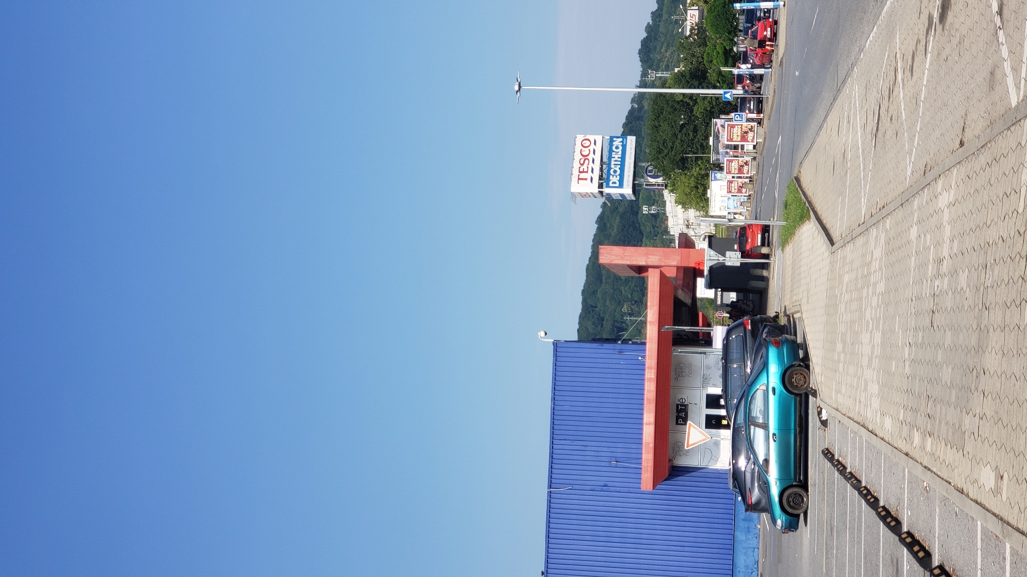 The image depicts an outdoor scene in the city of Prešov, Slovakia, likely a commercial area. In the foreground, a paved parking lot with white parking lines and concrete curbs is visible. Several cars are parked in the lot, including a teal sedan, a dark SUV, and another dark vehicle. The cars are positioned near a blue building with a red awning. Further back, a road runs alongside the parking lot. On the road, several cars are driving or parked, including red and black vehicles. A large billboard displaying "TESCO" and "DECATHLON" is visible in the distance, indicating a commercial shopping area. The sky is clear and blue, and a lush green hillside with trees forms the background. The scene appears to be set during daytime with bright sunlight.