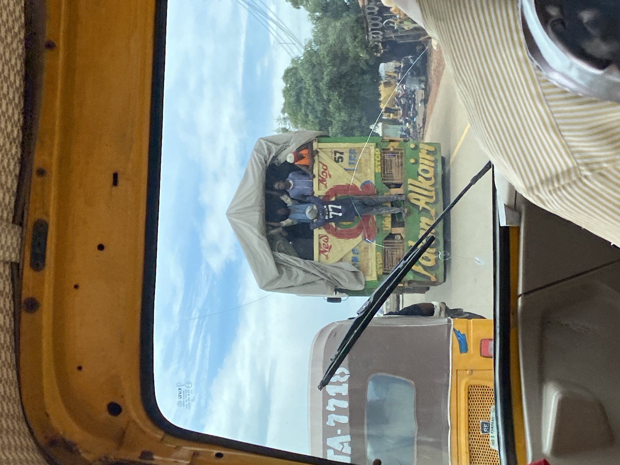 The image, taken from inside a vehicle, shows a street scene in Rafin Par, Nigeria. In the foreground, a portion of the interior of the vehicle is visible, including a yellow dashboard and a person wearing a horizontally striped yellow and white shirt. The windshield offers a view of the road ahead, where a brown and yellow auto-rickshaw with license plate "TA-7718" is driving to the left.
To the right of the auto-rickshaw, a large, green truck is driving in the same direction. The truck is covered with a light-colored tarp, and several people are standing on top of it, some visible through the gaps in the tarp. One person is wearing a blue shirt with the number "77" on the back, and another is wearing an orange vest. The truck has writing on its side, including "Mod" and "57", and what appears to be a name in yellow lettering, "Alkaliri". The truck also has colorful circular graphics and a "STOP" sign.
The environment appears to be an urban or semi-urban area, with trees and buildings visible in the background. Some motorcycles and other debris are present on the side of the road. The sky is cloudy. The overall impression is a busy street with traffic and people engaged in transport.