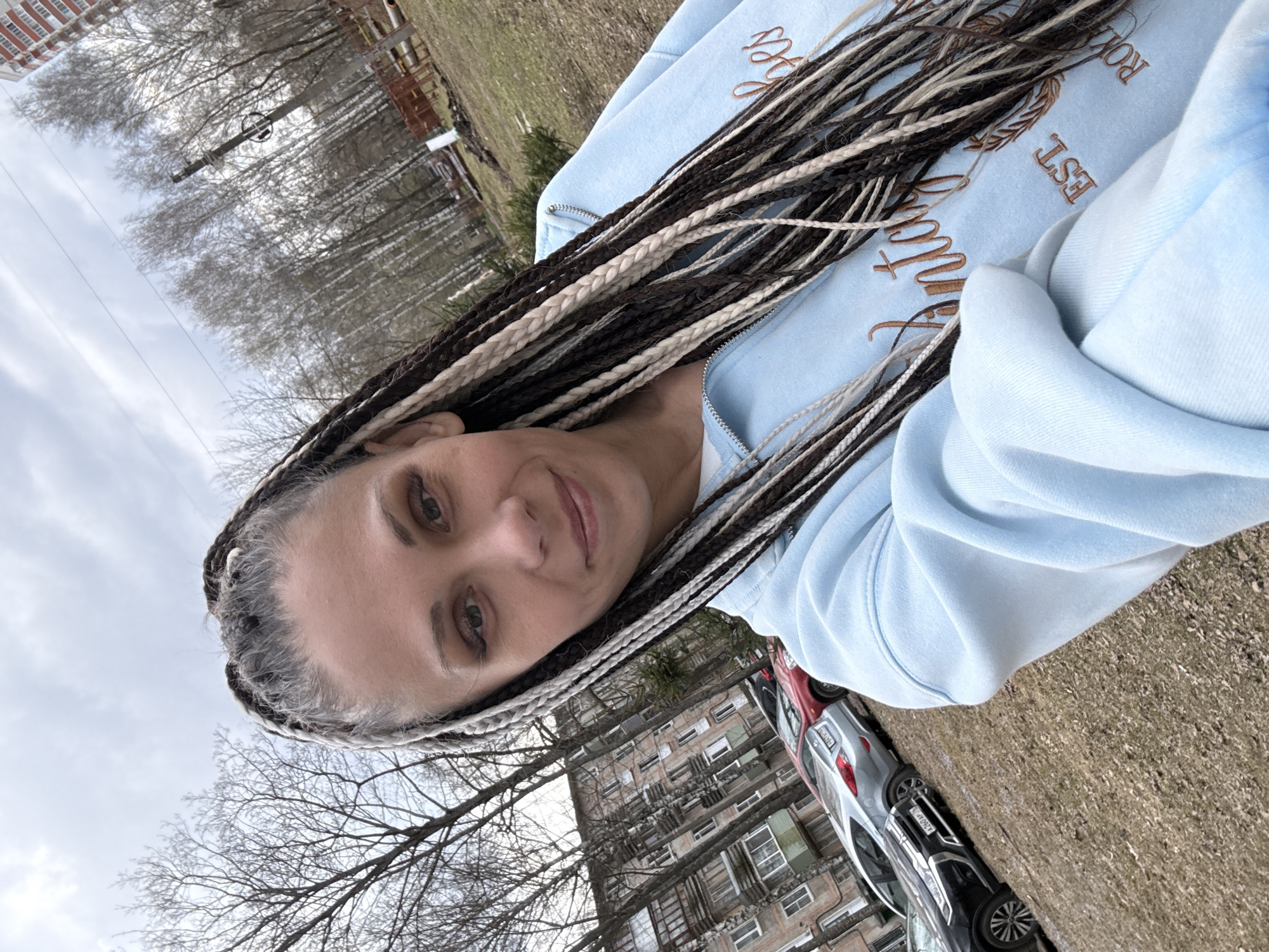 The image shows a woman taking a selfie outdoors in a residential area, likely in Rusanovo, Russia. She is positioned slightly off-center, looking directly at the camera with a calm, subtle smile. Her eyes are light-colored. She wears a light blue zip-up hoodie, which features embroidered text in a brown-gold thread; partially visible words include "EST." and what appears to be parts of "ROMAN" or "Saint Anthony". Her hair is styled in long, numerous braids, incorporating dark brown, light blonde/white, and grey strands.
The background reveals an urban or suburban setting. Several cars are parked on a patch of dry ground with sparse grass, including silver, black, and red vehicles, with Russian license plates partially visible (e.g., "M260 AP 70"). A large, multi-story apartment building with numerous windows is prominent behind the cars. Beyond this, many bare deciduous trees, possibly birches, line the horizon under an overcast sky. The scene depicts a cool, daytime setting, likely late winter or early spring, characterized by the leafless trees and diffused light.
