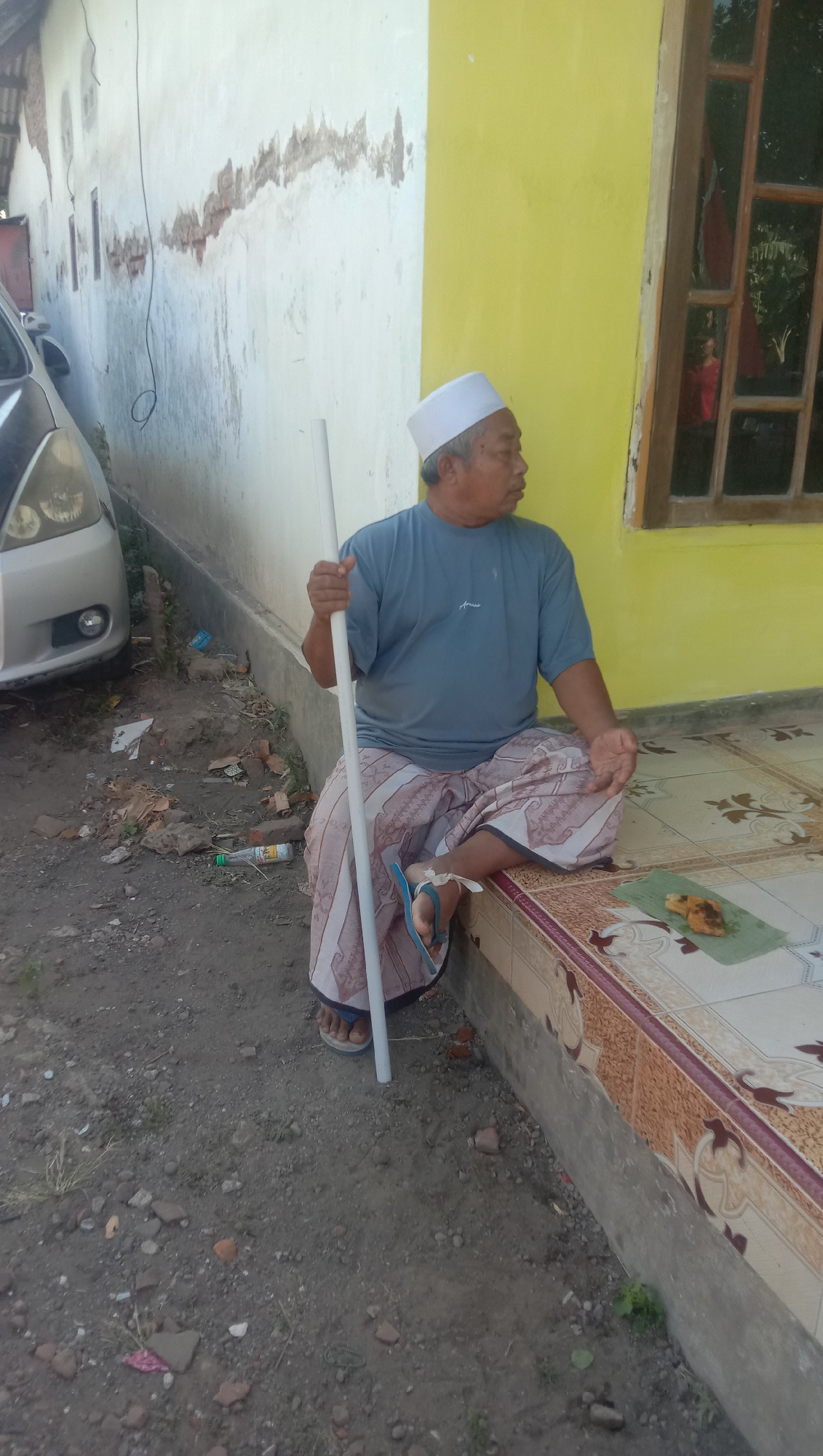 An elderly Indonesian man wearing a white peci and a blue t-shirt is seated on a tiled porch in front of a yellow and white building. He is holding a white pole with his right hand and appears to be looking to his right. His legs are crossed, and he is wearing a patterned sarong and blue flip-flops. A portion of a silver car is visible on the left side of the image. The ground is dirt and gravel, with some scattered debris. A window with a red curtain is visible in the yellow wall of the building. A banana leaf with food on it is placed on the tiled porch to the man's right. The scene appears to be located outdoors in what seems to be a residential area of Ngempit, Indonesia.