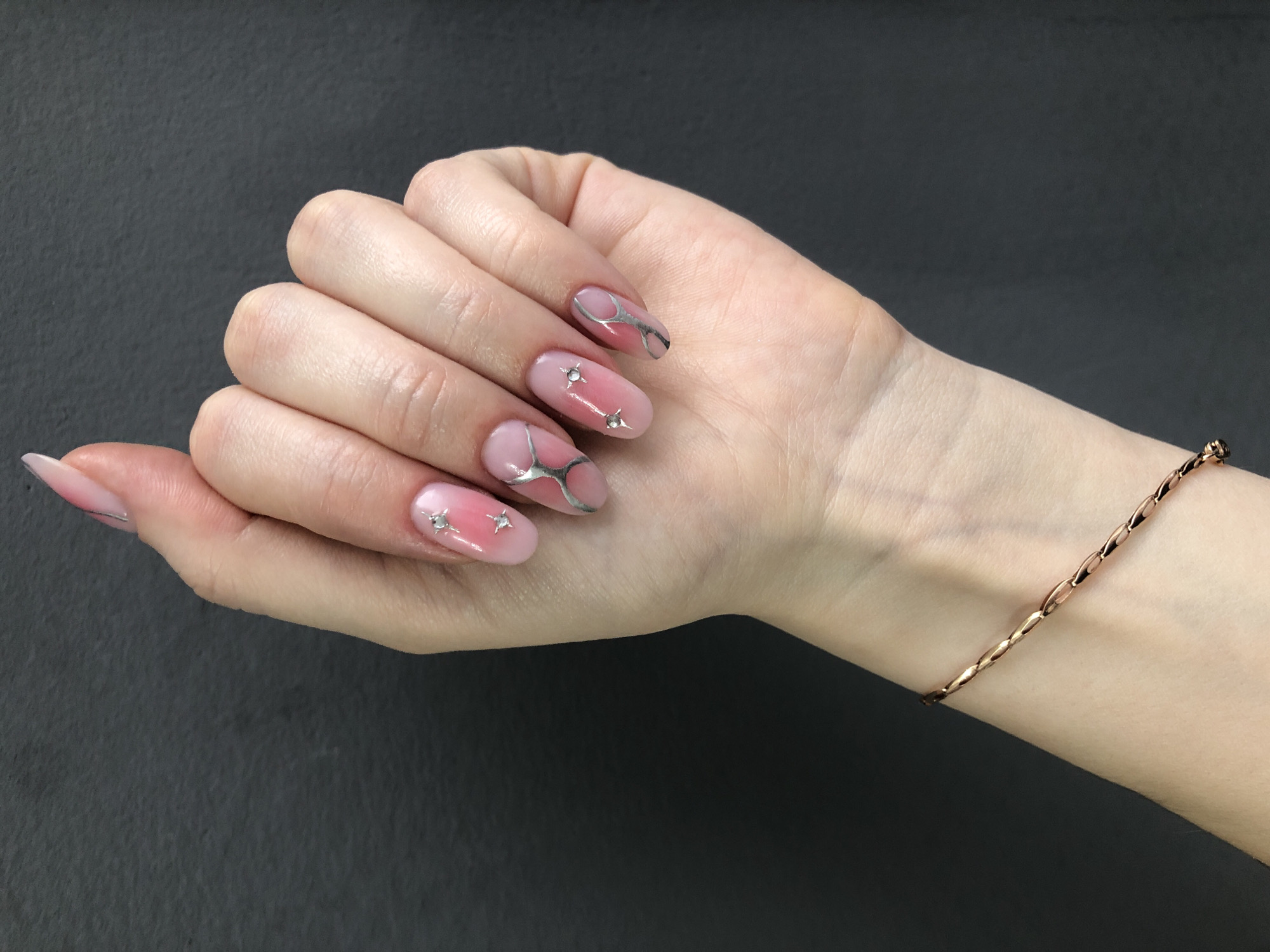 This close-up shot, taken in Chaikovsky, Russia, features a person's left hand displaying elaborately manicured nails against a dark, textured background. The hand is positioned with fingers slightly splayed and curved, showcasing the nail art. The nails are long, with an oval or almond shape, and feature a gradient effect with a light pink color on the nail bed blending into a sheer or clear tip.
Each nail is adorned with unique silver, metallic, abstract designs that appear to be hand-painted, curving and swirling across the nail surface. The ring and middle fingers also feature small, clear, star-shaped or spike-shaped studs embedded in the polish. A thin, rose gold or copper-colored chain bracelet, composed of small, elongated, flat links, is visible on the wrist. The lighting is even, suggesting an indoor setting. There is no discernible activity other than presenting the nails. No visible text is present. The overall image emphasizes detailed nail art and jewelry as fashion accessories.