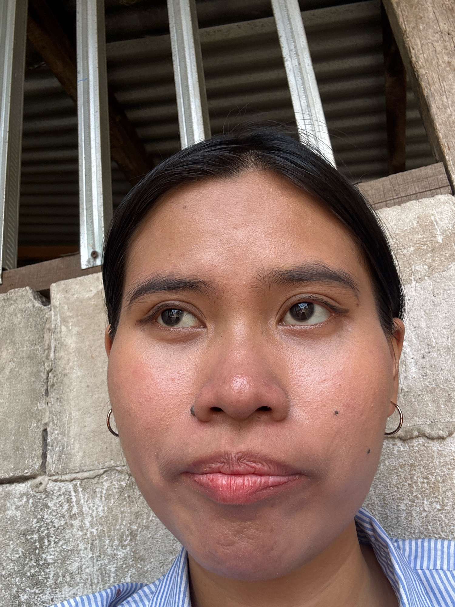 A close-up, medium shot photograph features a young woman's face against a textured gray concrete wall and a corrugated metal roof structure. The woman, with dark hair pulled back, is positioned in the center, looking slightly to her left with her lips pursed. Her skin appears slightly shiny, with visible pores and two small moles on her right cheek. She wears a blue and white striped shirt, and a silver hoop earring is visible on her left earlobe. The background consists of a weathered wooden plank and vertical metal beams supporting the corrugated roof, suggesting an outdoor, possibly industrial or construction site setting in East Jakarta, Indonesia.