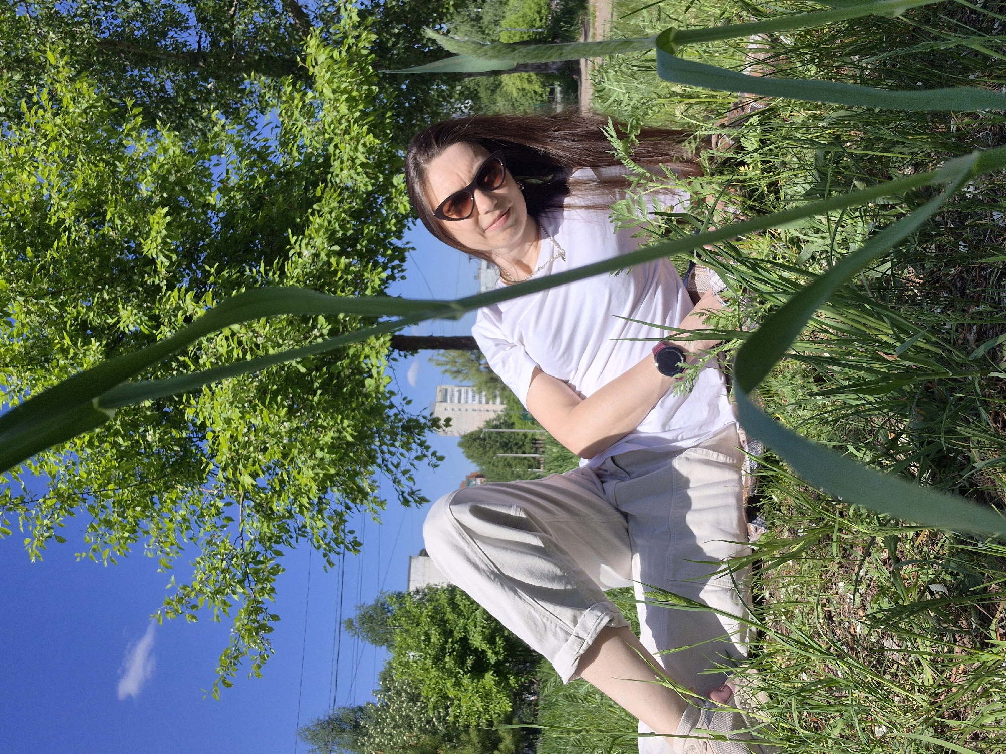 A woman with long dark hair and sunglasses is sitting in a grassy area. She is wearing a white t-shirt and light-colored pants. The woman is looking at the camera. A tree is to the left of the woman and some tall grass surrounds her. A building is visible in the background and the sky is blue with some clouds. Electrical lines are visible above. The location is Unknown, Unknown.