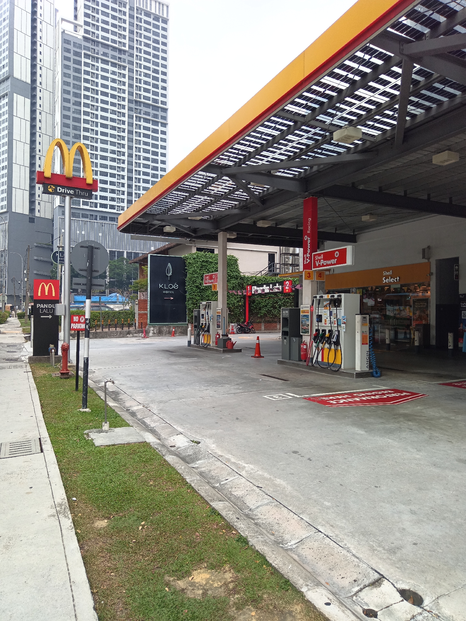 In the city of Kuala Lumpur, Malaysia, a Shell petrol station is captured with a McDonald's restaurant and high-rise buildings in the background. The petrol station features a covered area with solar panels on its roof, and several fuel pumps are visible. Red and yellow branding is prominent on the station. A McDonald's Drive-Thru sign is positioned on the left side of the image, near two vertical black signs, one with a McDonald's logo and "Pandu Lalu" text, and another displaying "NO PARKING." Across the street, a hotel named "KLOE HOTEL" is advertised on a large black sign. The ground is paved, with some grass and drainage systems present. The sky is overcast.