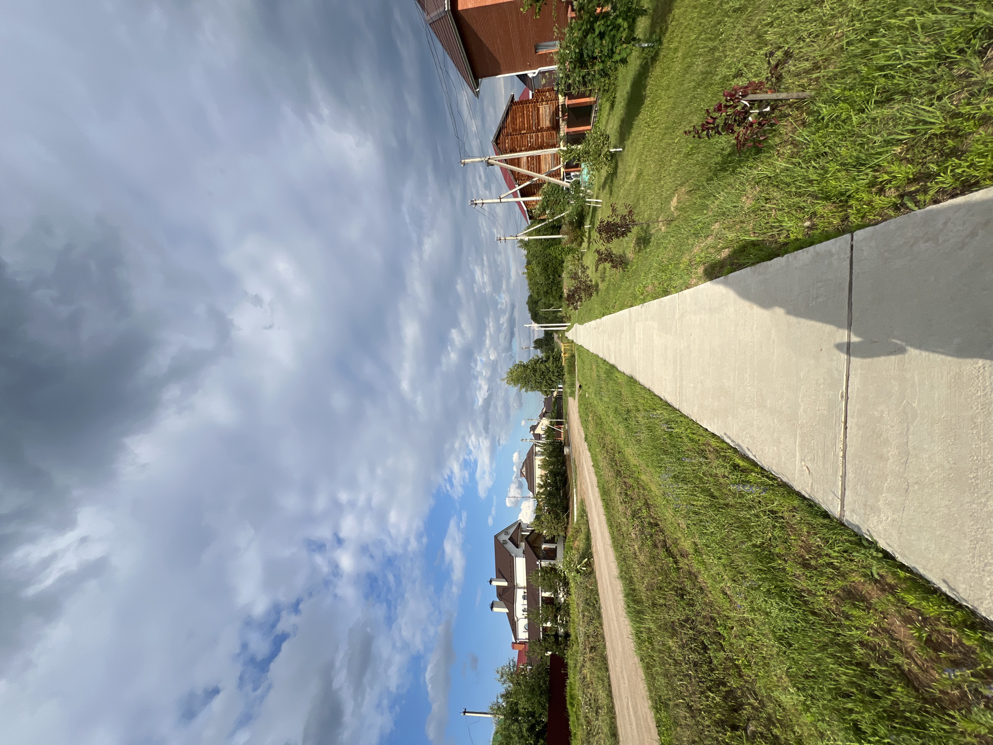 The image depicts a straight, paved sidewalk running through a suburban area with houses and greenery. The sidewalk is made of concrete slabs and is bordered by grassy areas. On the left side of the sidewalk, a dirt road is visible, with houses and trees beyond it. On the right side, there are more houses, fences, trees, and saplings planted in the grass. Power lines with wooden poles are also present throughout the scene. The sky is cloudy with patches of blue sky visible. The overall impression is of a quiet, residential street in a rural or suburban setting. The location is identified as Noviy, Russia. There are no people visibly present in the image, but a shadow of a person is cast on the sidewalk from the foreground.
