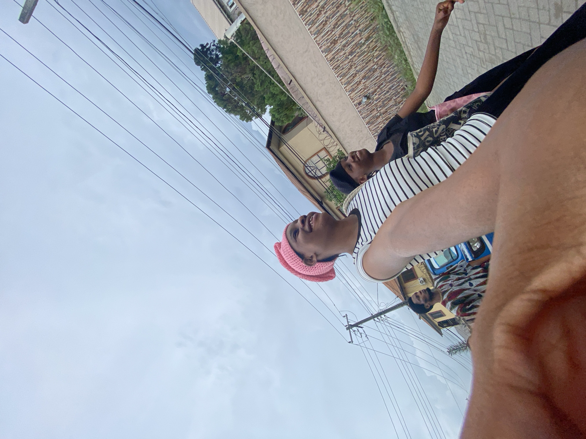The image depicts two individuals in an outdoor setting in Unknown, Unknown. One person, seen in the foreground, is wearing a pink hat and a striped shirt. The other person, slightly behind and to the side, is wearing a black shirt. The background includes buildings, trees, power lines, and a cloudy sky.