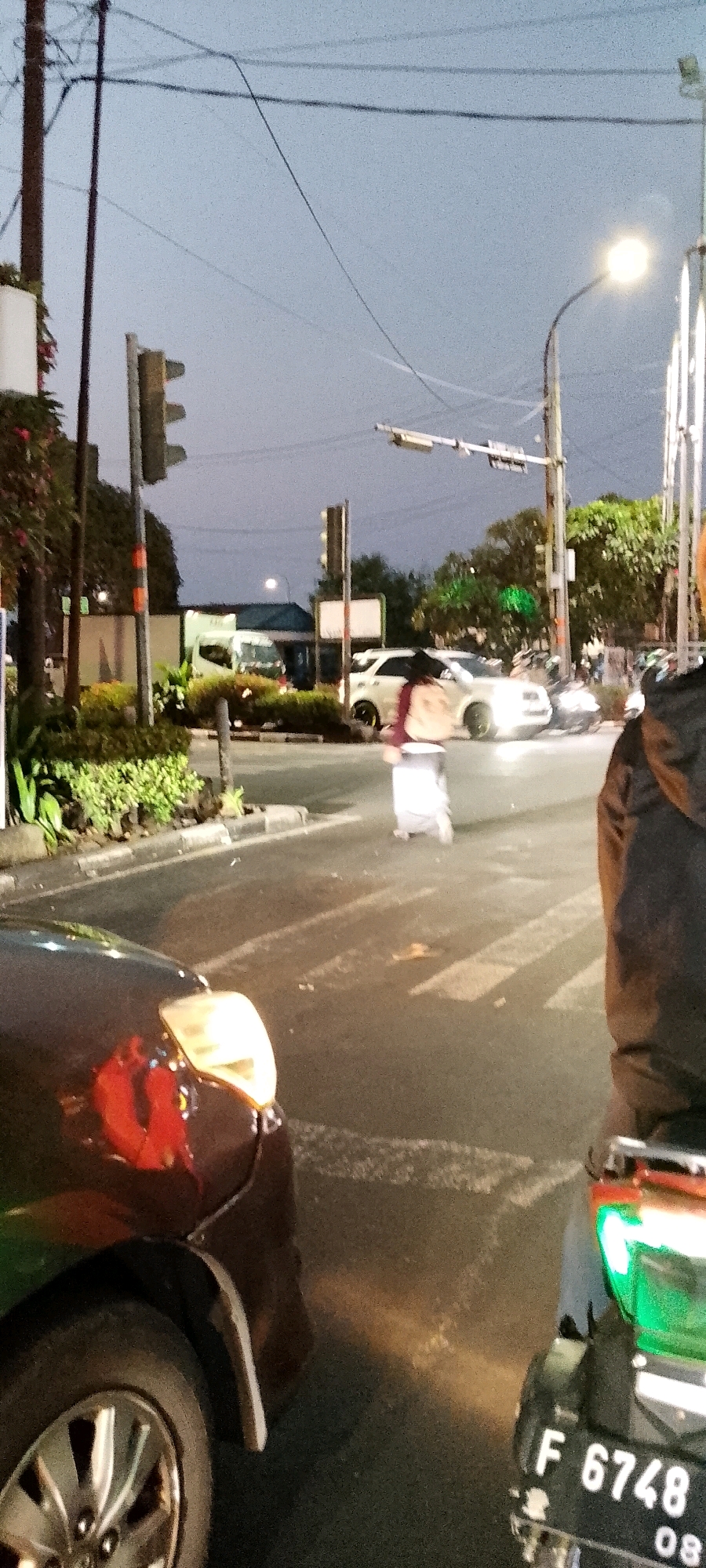 The image captures a street scene at dusk in Depok, Indonesia. In the foreground, a dark-colored car is parked on the left side of the frame, its headlights illuminated. To the right, a motorcycle is also parked, with its license plate reading "F 6748 08" visible.
Further down the street, a white SUV is stopped at a pedestrian crossing. A person dressed in white, possibly a pedestrian, is walking across the street. Traffic signals are visible on poles throughout the scene, indicating a road intersection. Trees and foliage line the side of the road, and overhead power lines stretch across the sky. The overall atmosphere suggests a typical urban environment in Indonesia during the evening.
