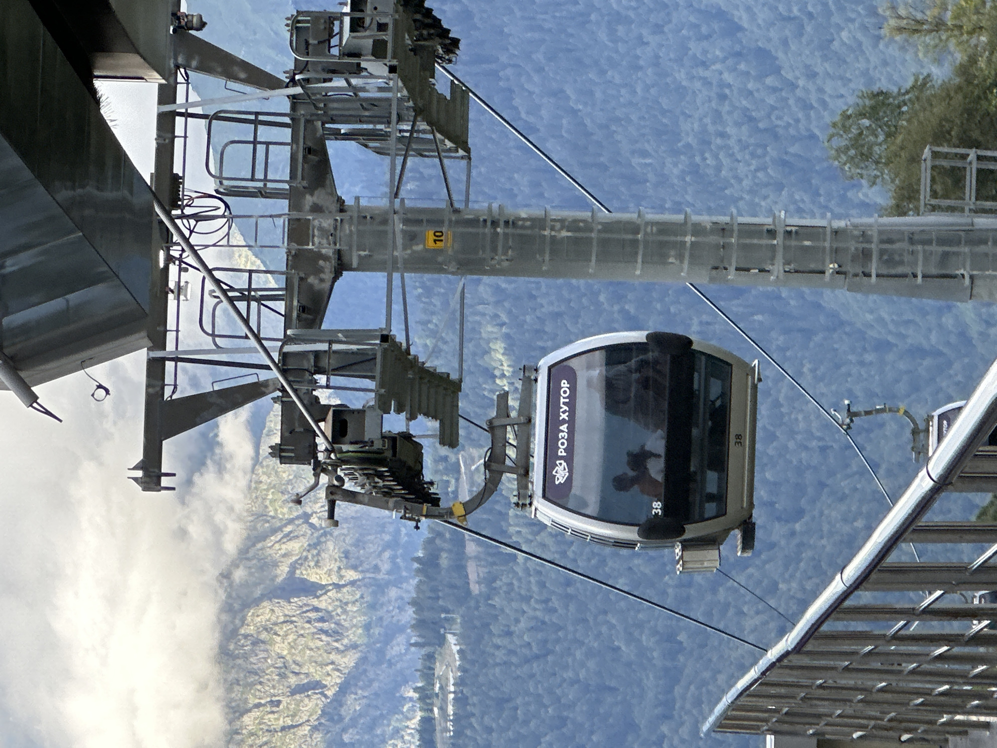 A cable car travels upwards in the mountain resort of Rosa Khutor in Estosadok, Russia. The cable car is white with purple lettering that reads "РОЗА ХУТОР" and the number "38" on the exterior. Inside the cable car, two passengers are visible. The cable car is suspended from a large metal tower that supports the cable. The background features a densely forested mountain slope with some rocky outcrops and a cloudy sky. A metal railing and walkway are visible in the foreground.